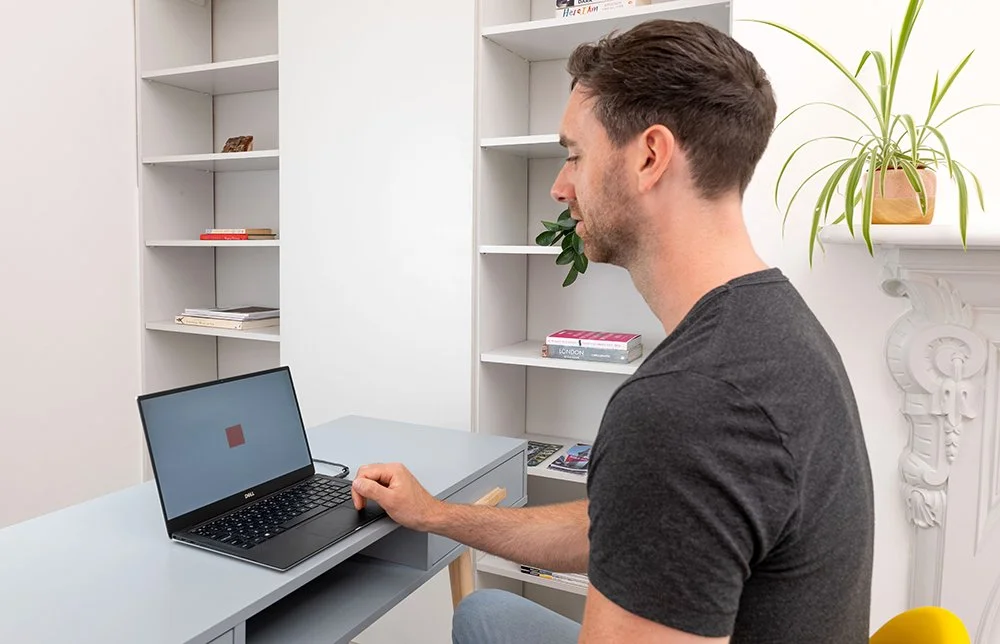 A man using a laptop at a light blue desk in a white room with bookshelves and a potted plant.