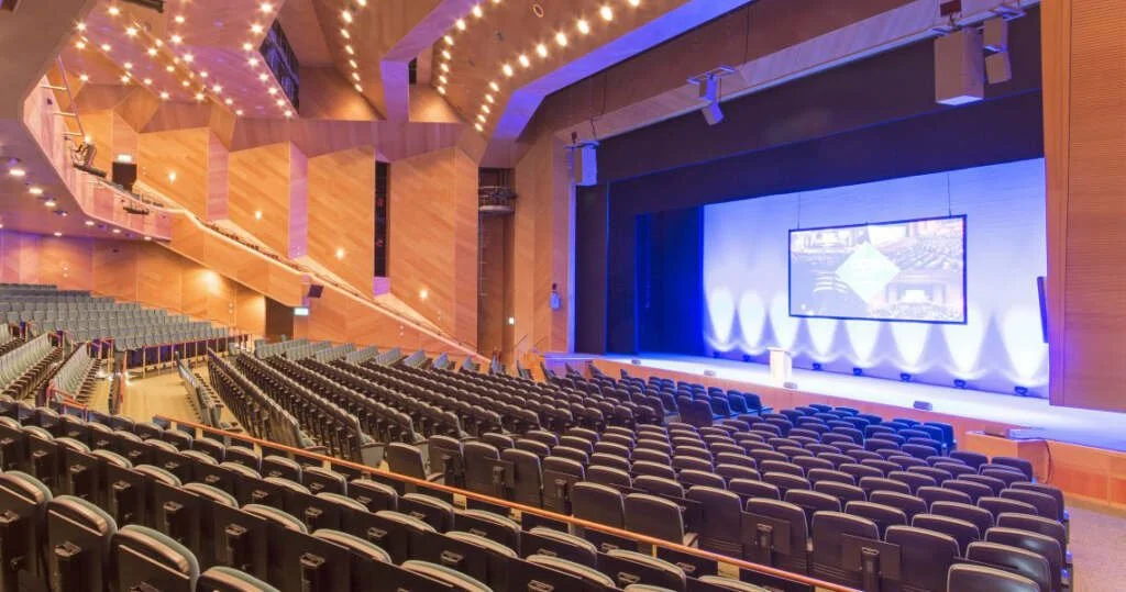 A large audience of women seated in a conference hall with blue stage lighting and smoke effects.