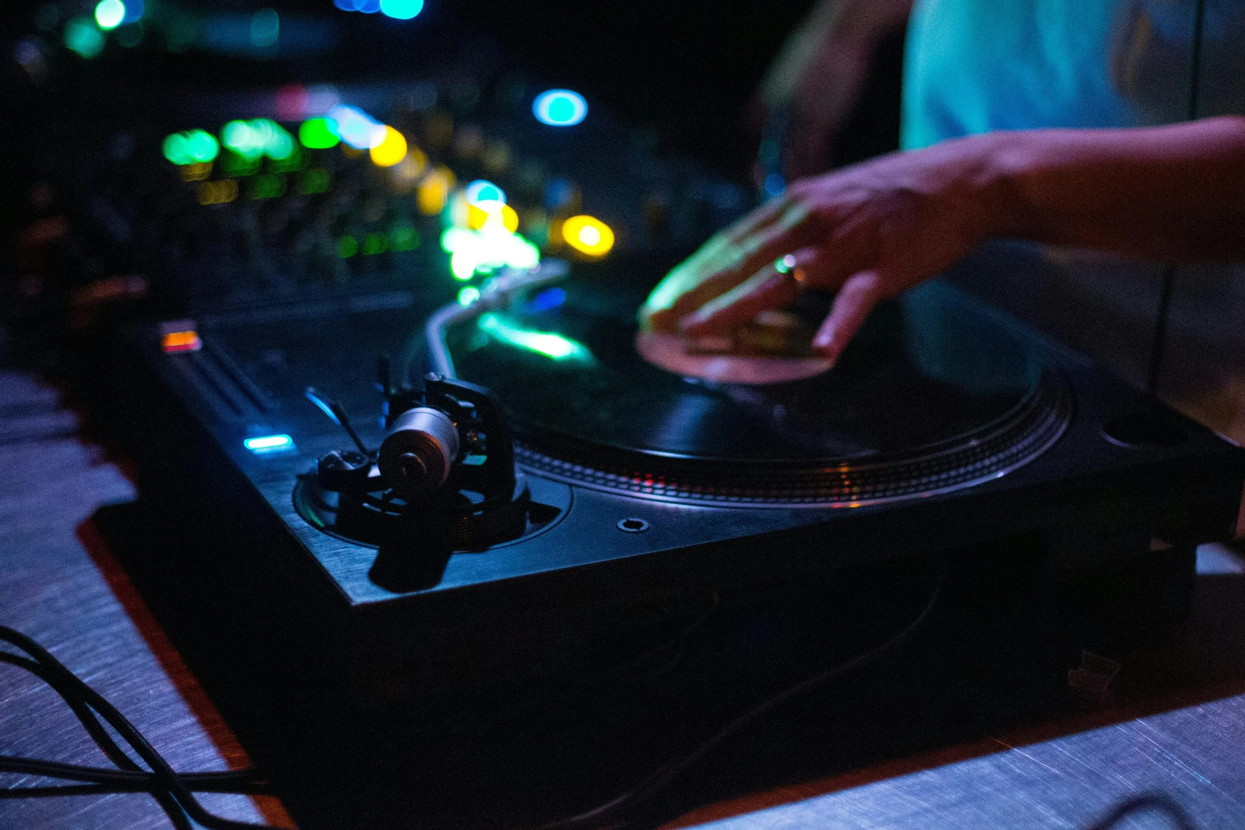 A DJ's hand moving a vinyl record on a turntable with colorful blurred lights in the background.