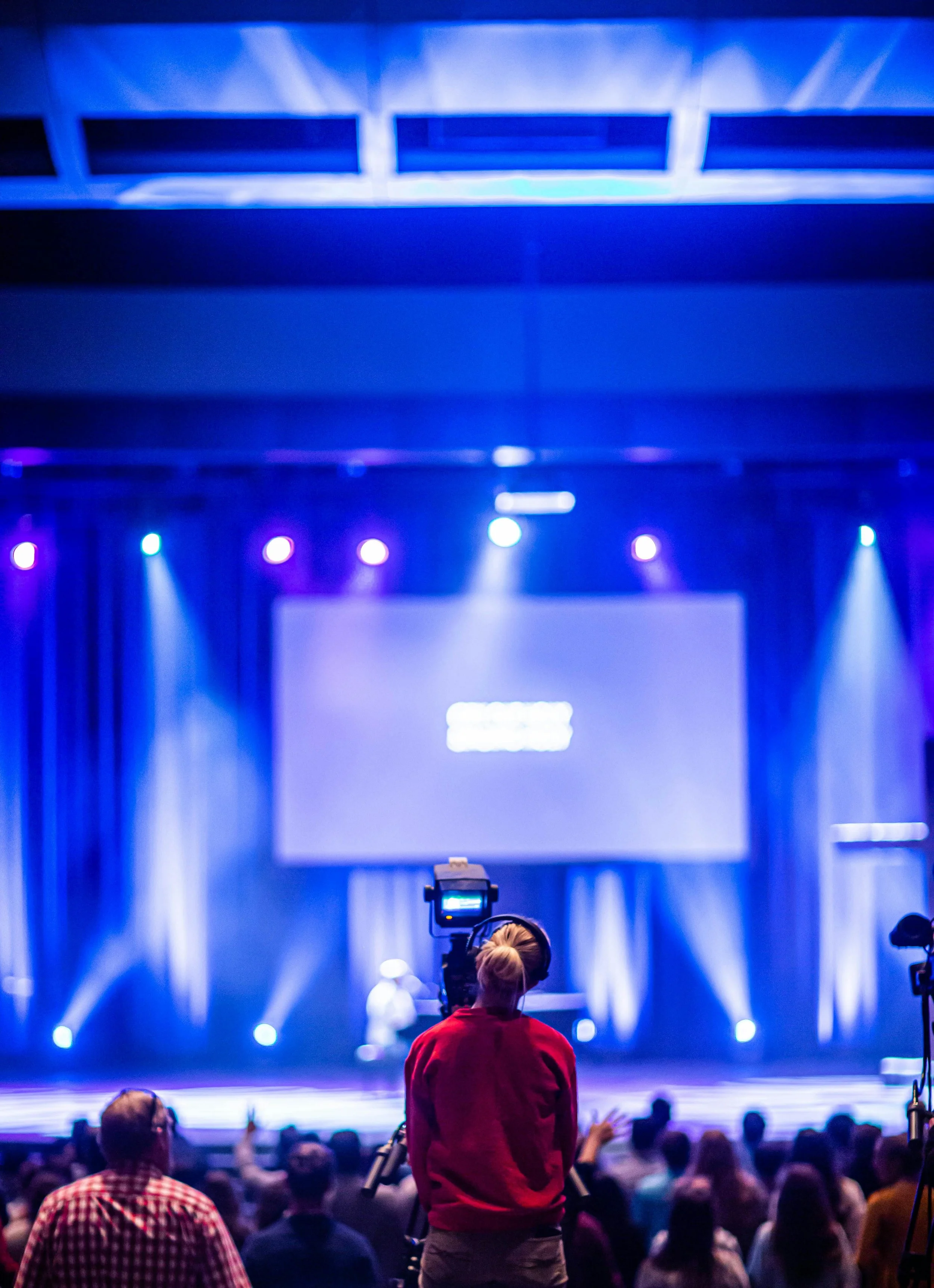 A person operating a camera on a tripod during a concert or event with blue stage lighting and an audience in front of the stage.