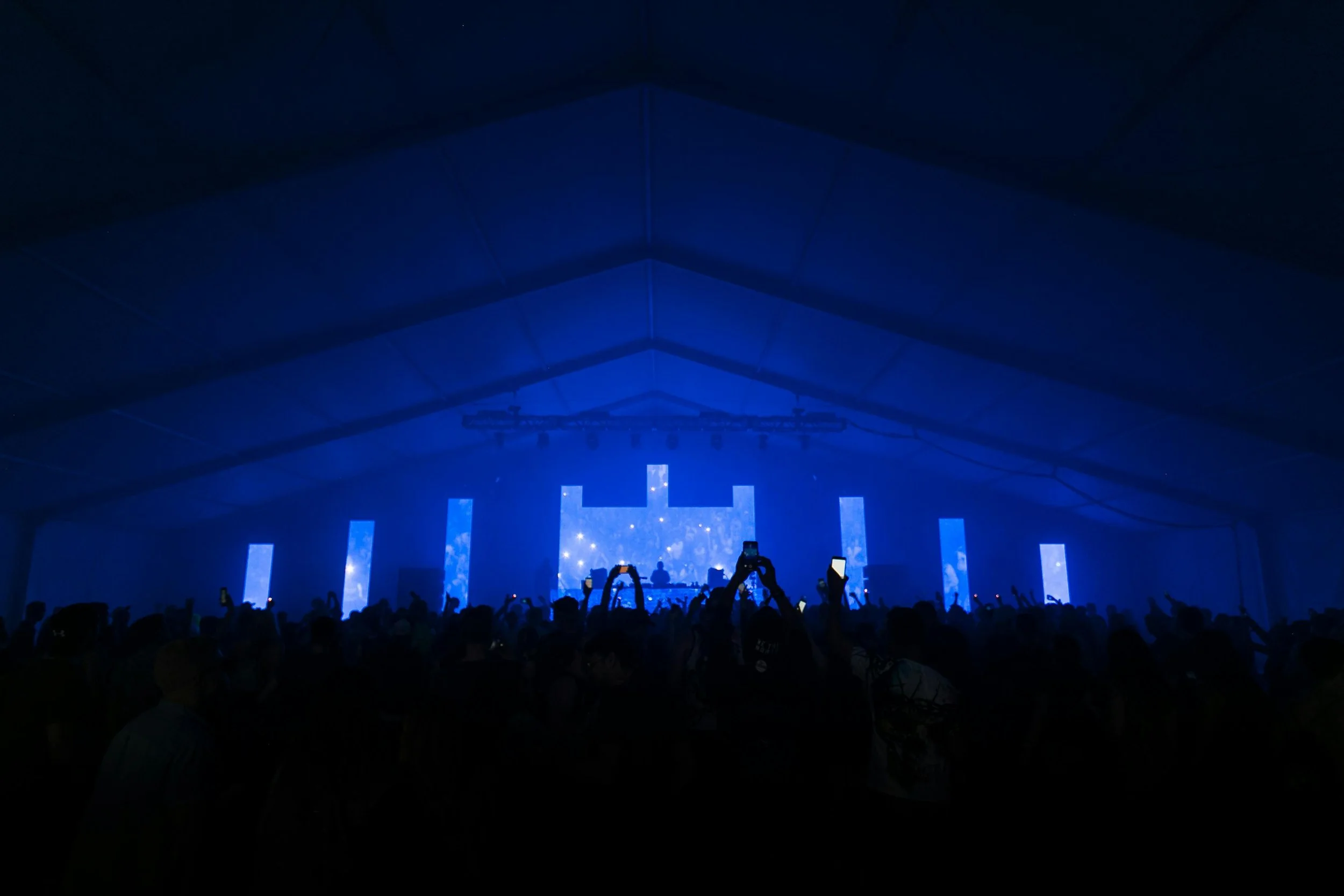 A crowded concert with a DJ on stage, blue lighting, and many people taking photos or videos with their phones.