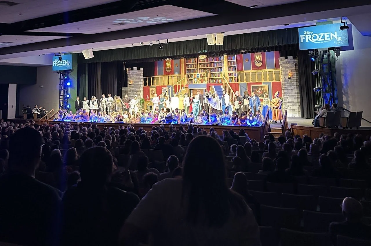 A stage production of Disney's Frozen Jr. with actors dressed in costumes, set against a colorful backdrop resembling a library or castle interior, with audience members watching attentively.