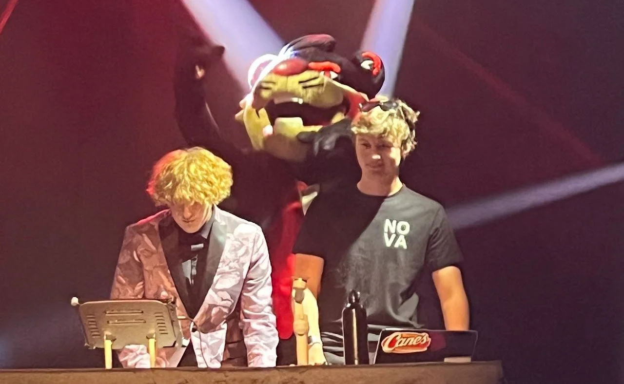 Two young men performing on stage with a large, colorful puppet of a creature with a big mouth and exaggerated features behind them. One man is wearing a pink patterned jacket, and the other is wearing a black NOVA T-shirt.