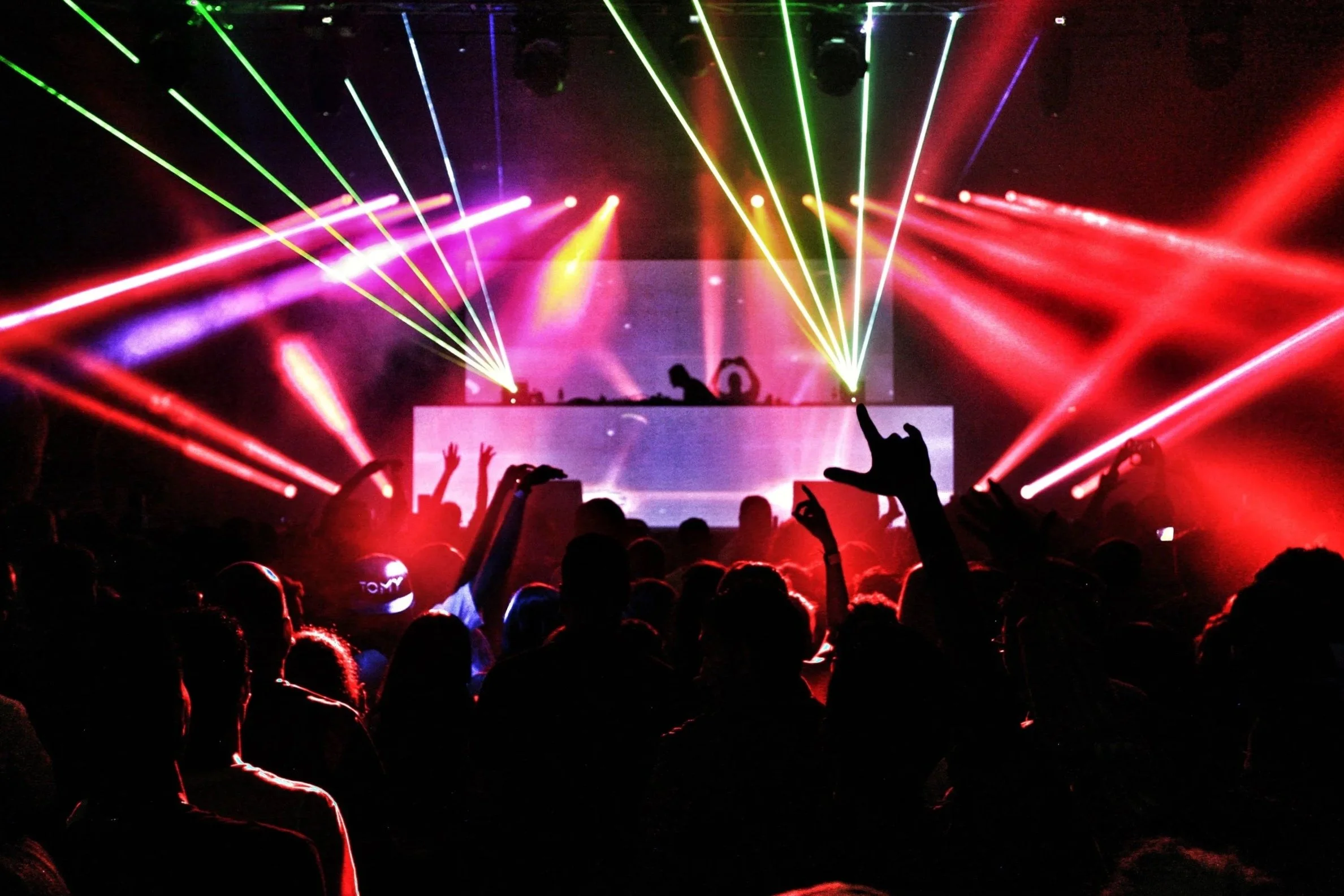 View from the DJ booth showing a DJ setup with controllers and a laptop, overlooking a crowded dance floor with party-goers under purple lighting.