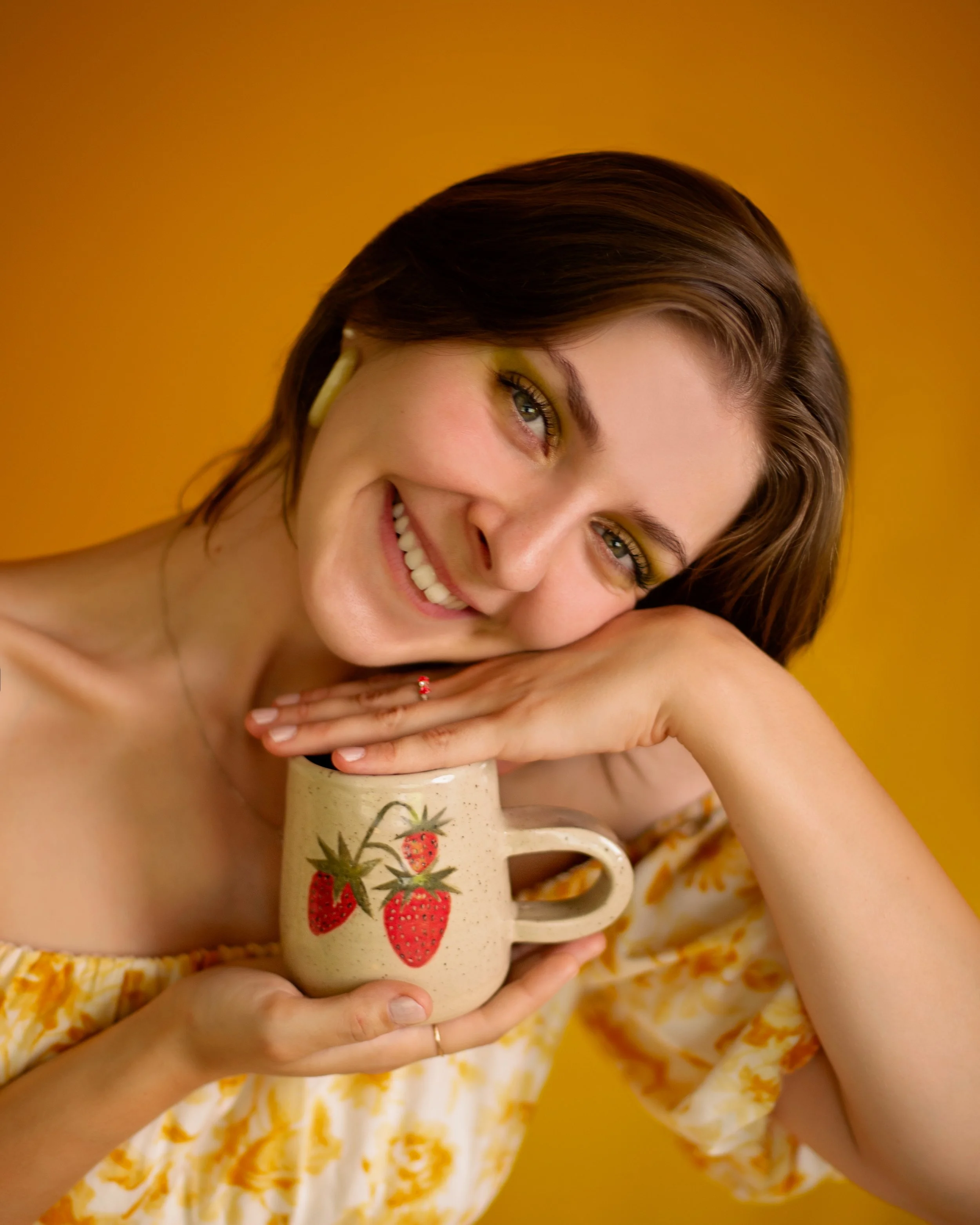 A woman with short brown hair and in a yellow floral dress, smiling and resting her head on a beige mug decorated with strawberries, against a yellow background.