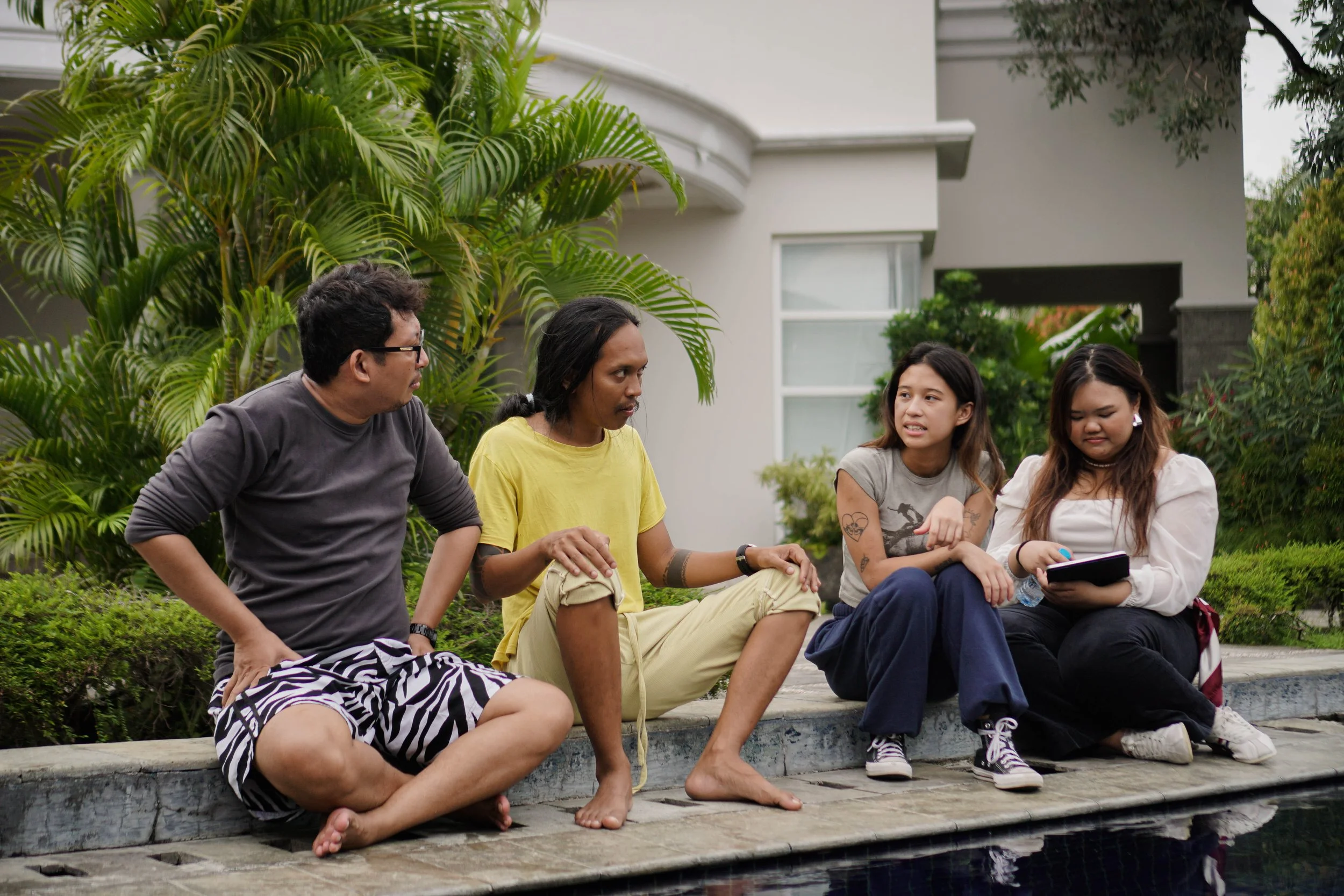 Four people sitting by a swimming pool having a conversation, with greenery and modern house in the background.