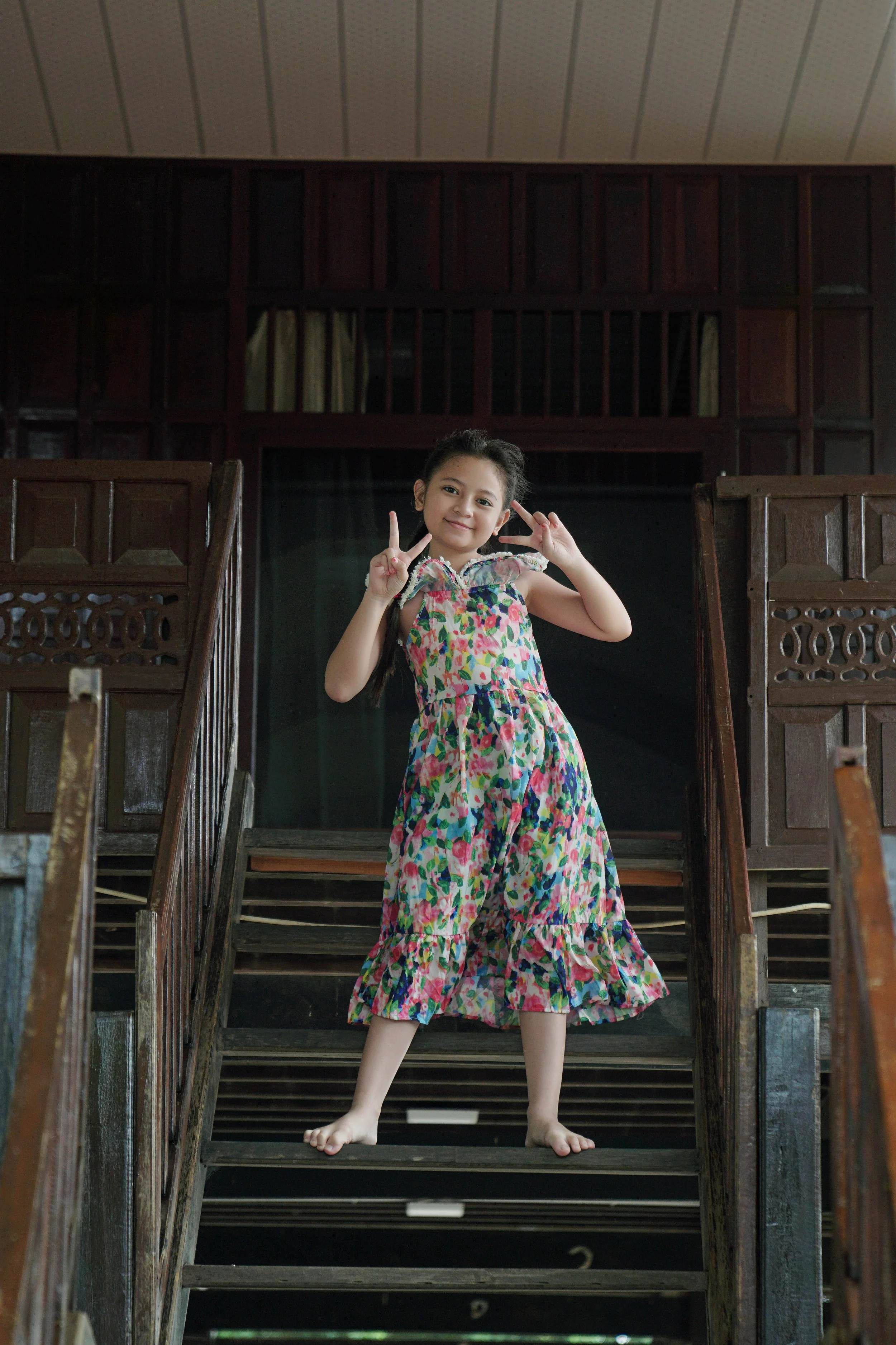 A young girl in a colorful floral dress stands barefoot on a wooden staircase inside a house, making peace signs with her hands and smiling.