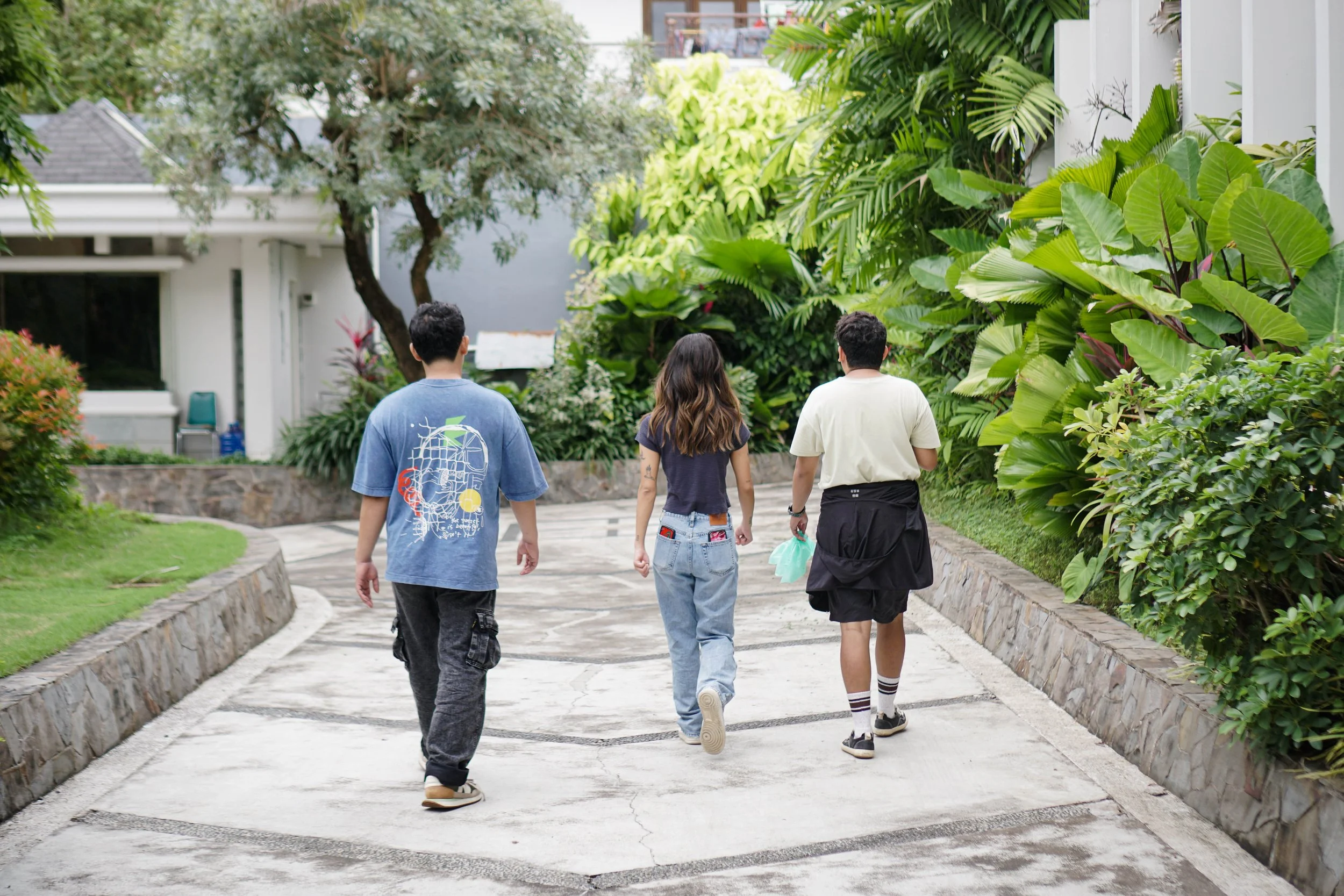 Three people walking down a paved pathway surrounded by lush greenery and trees in a residential area.
