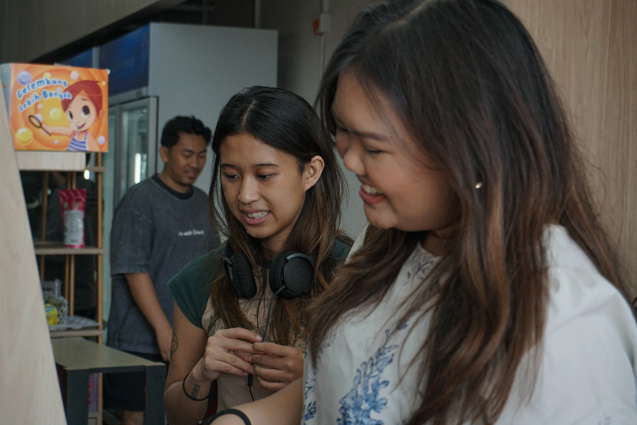 Two young women looking at something off-camera, one with headphones around her neck, smiling. A man is in the background with a shelf and a colorful sign with a cartoon girl holding a badminton racquet.