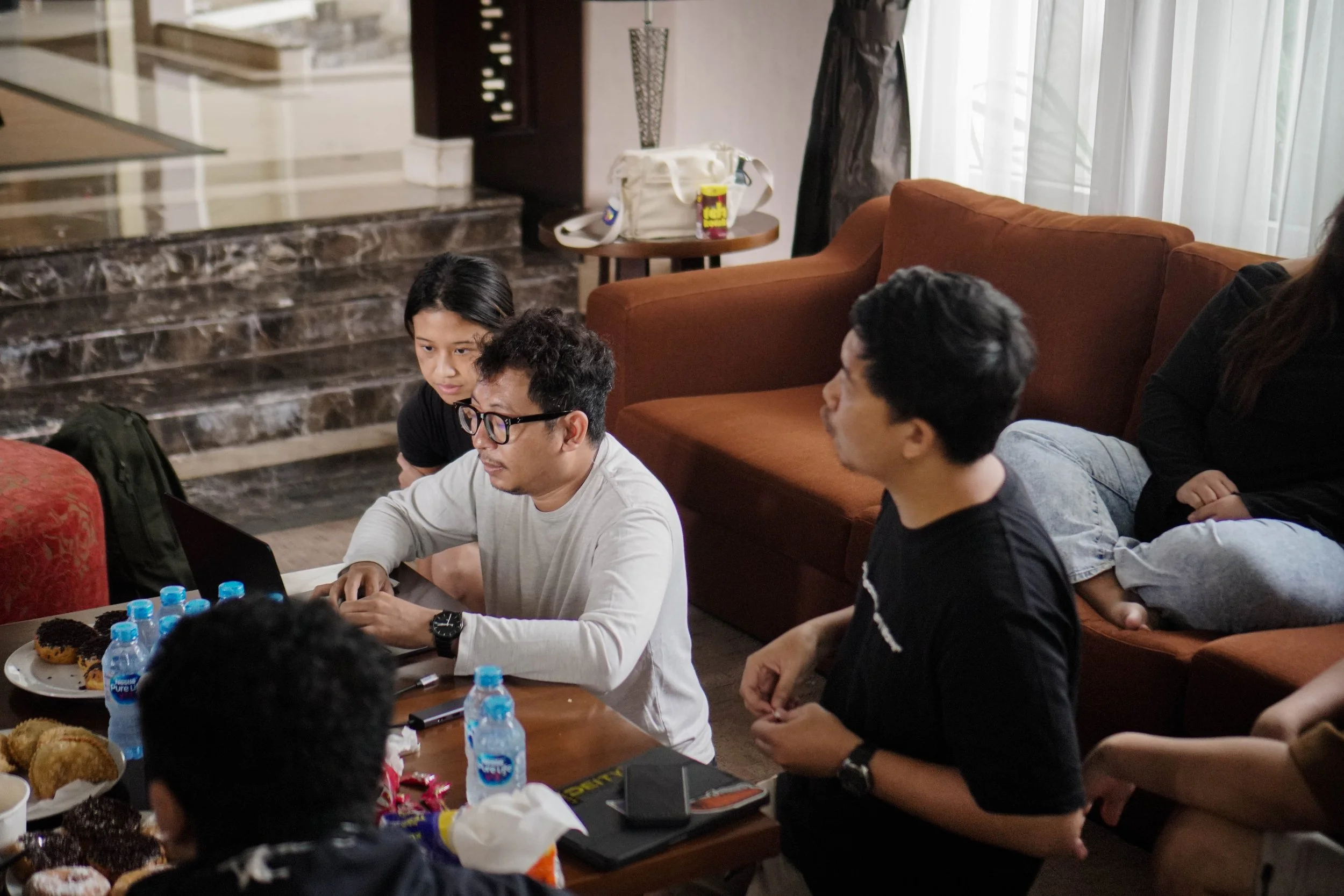 Group of people gathered around a table with food and water bottles, indoors in a living room setting.