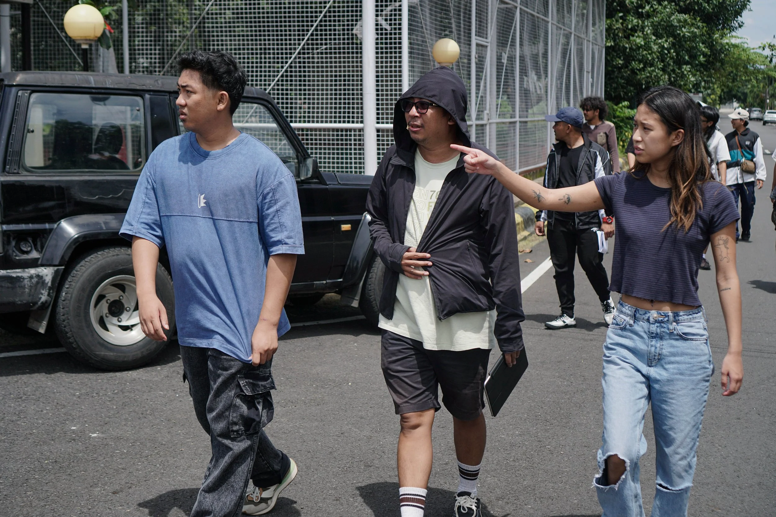 A group of young people walking on the street, with a woman pointing, and a black vehicle parked nearby.