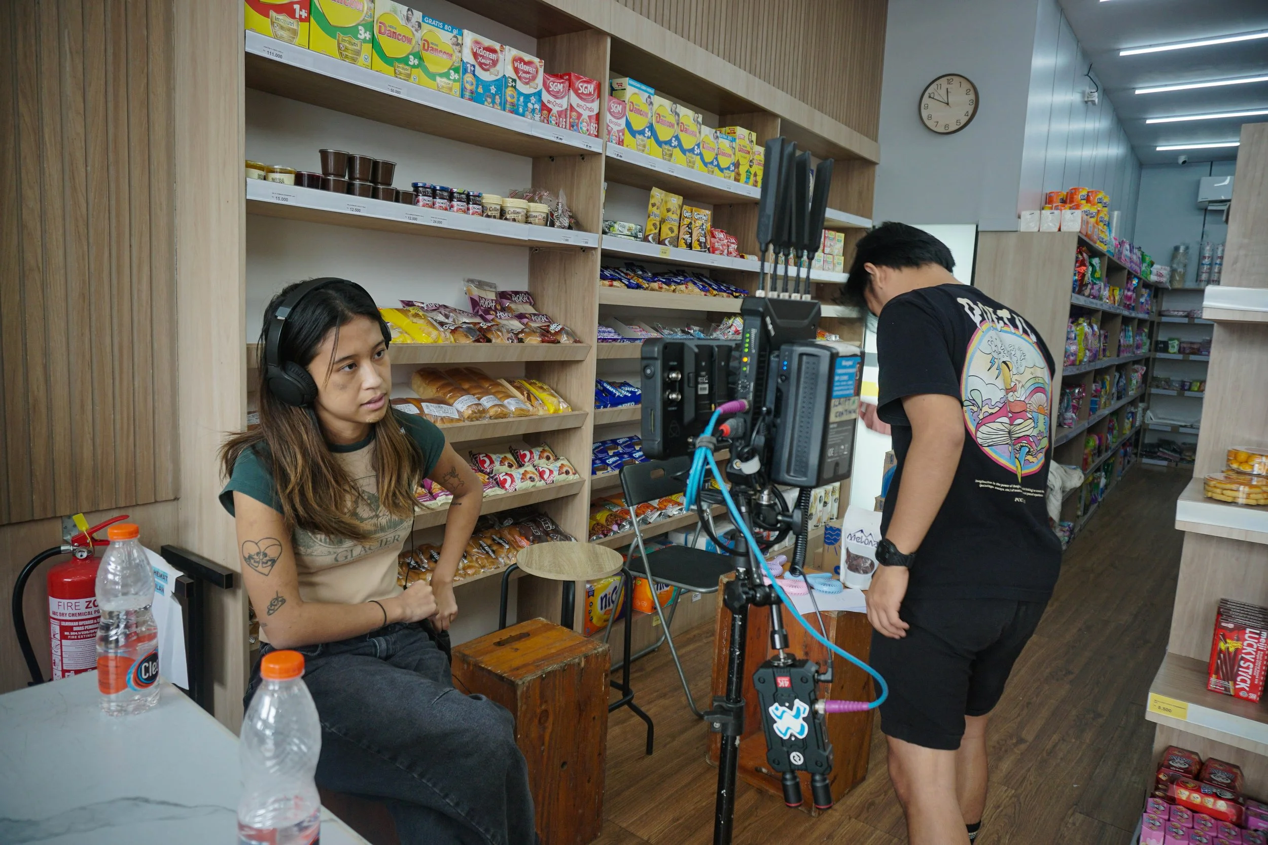 A woman wearing headphones sits in a store aisle with snack shelves behind her, while a man operates camera equipment, possibly filming or photographing her.