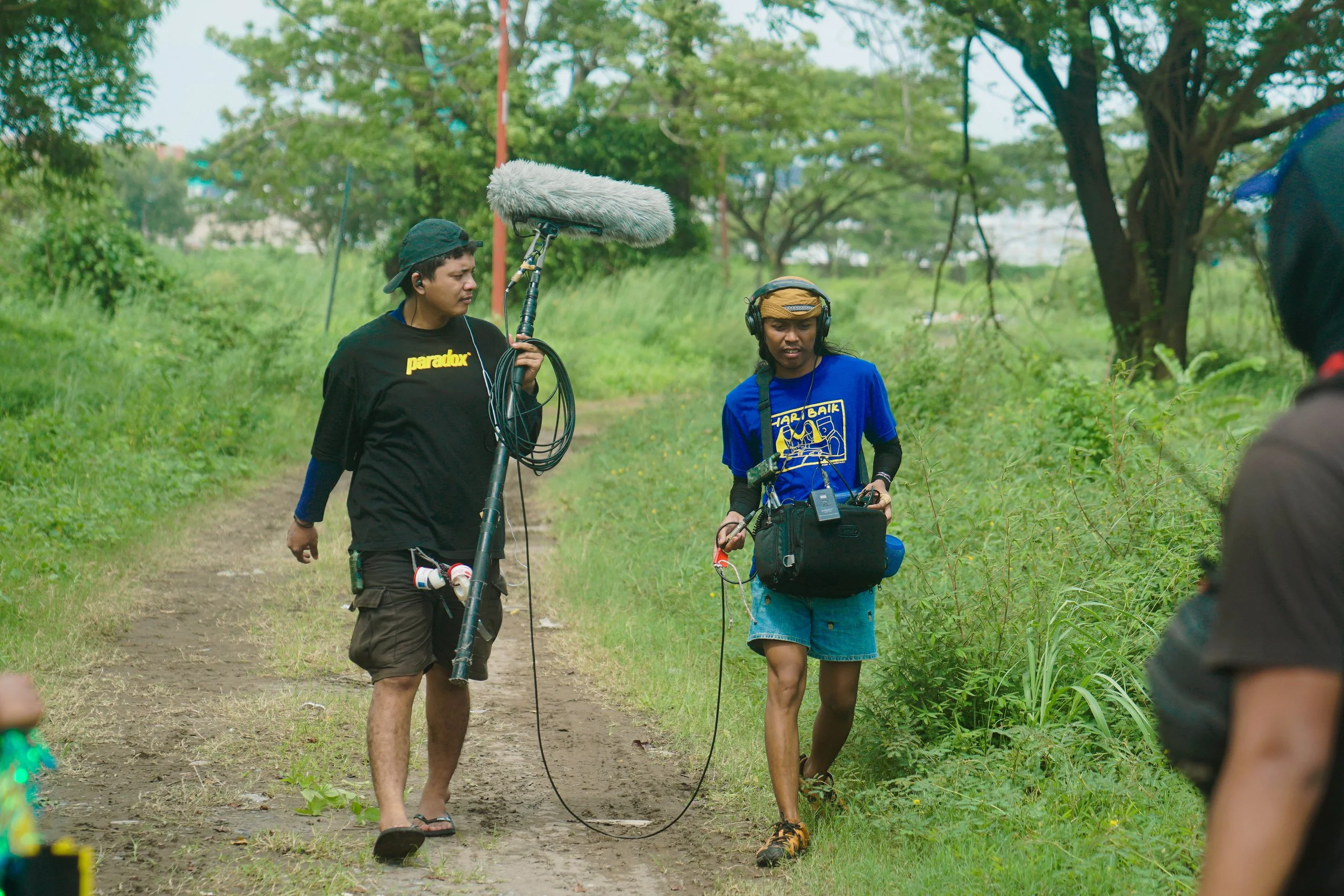 Film crew filming outdoors on a grassy dirt path with trees in the background. One person wearing a blue shirt and headphones, holding equipment, is being filmed by another crew member holding a boom microphone.