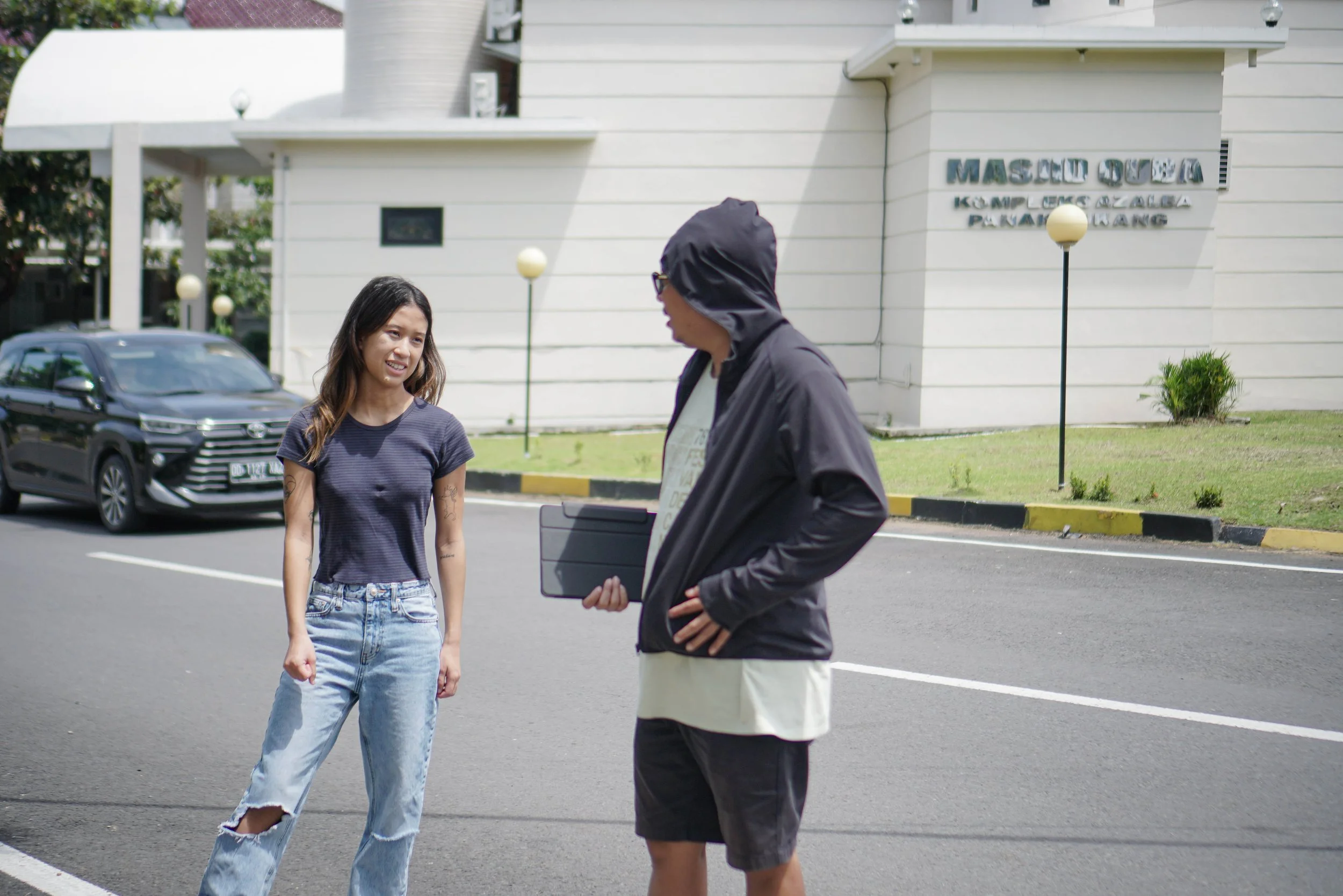 A woman talking to a man with a tablet in front of a white building with a sign that reads "MASJID" on the wall.