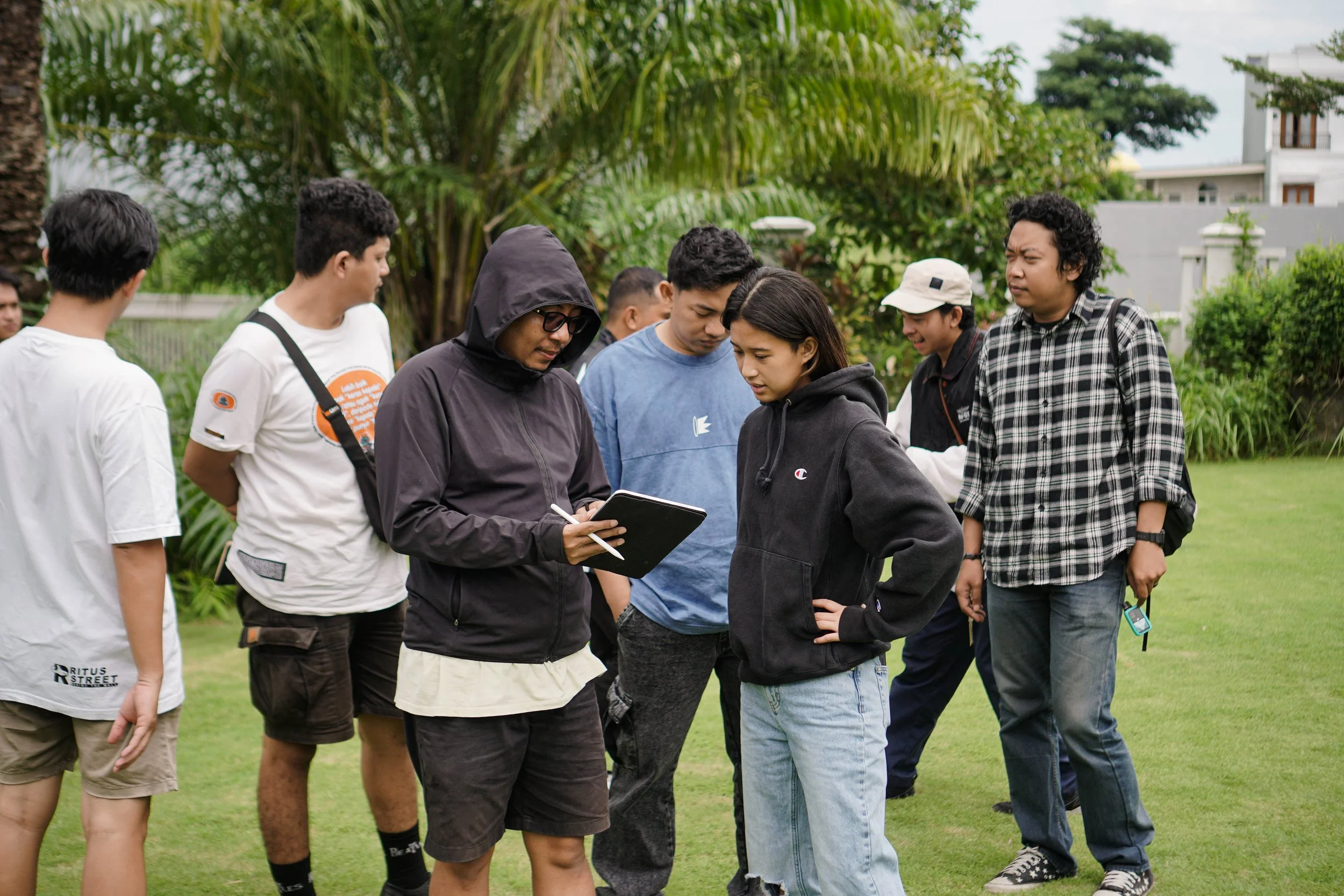 A group of young people standing outdoors on a grassy area with trees, engaged in a discussion or activity, with one person holding a clipboard and writing.