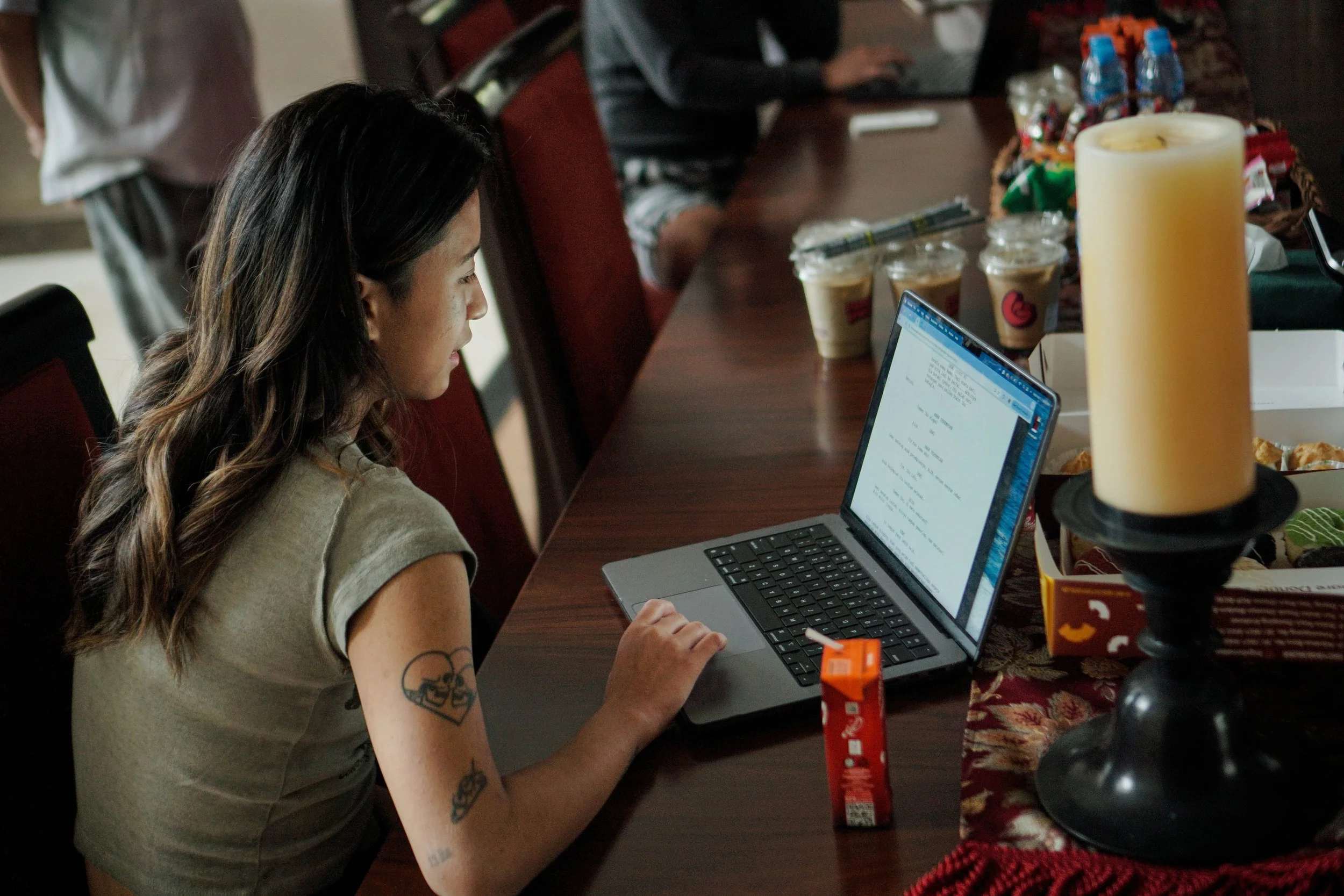 A woman with long dark hair and a tattoo on her arm is sitting at a dining table working on a laptop. on the table are several drinks, snacks, and a large candle. In the background, a person is using a tablet or phone, and another person is standing.