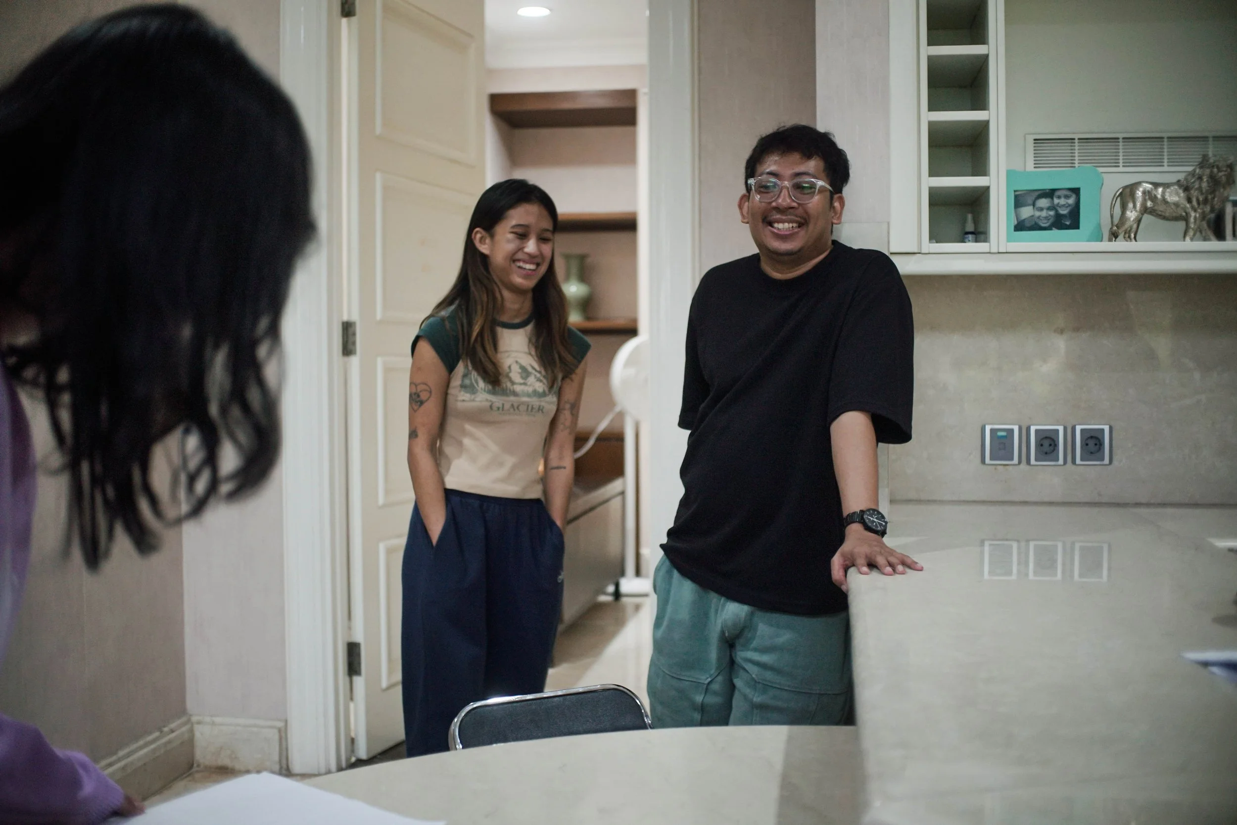 Three people in a kitchen laughing and smiling at each other, two women and one man, with one woman partly visible on the left.