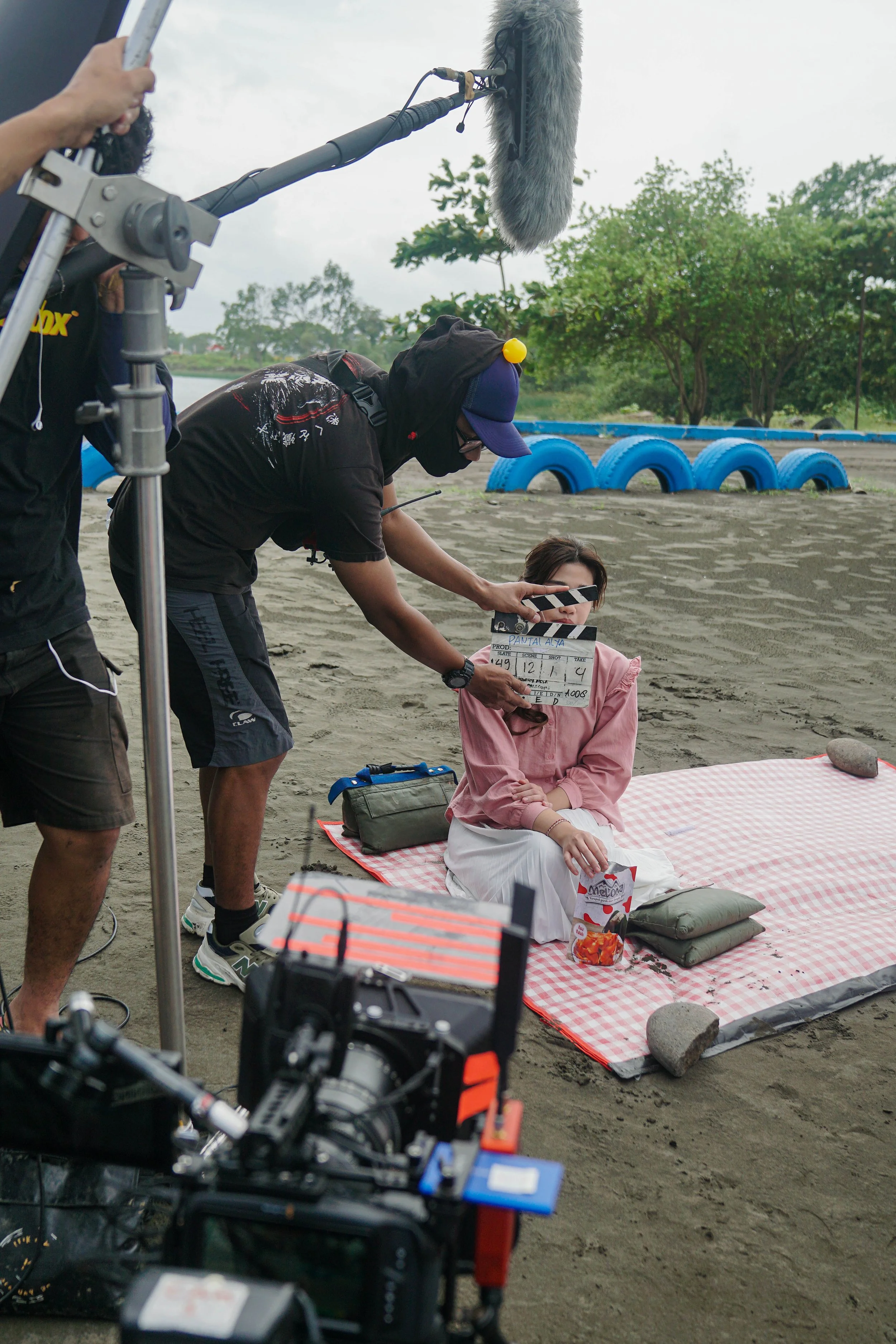 Film crew filming a woman sitting on a picnic blanket on a sandy area with trees in the background. The woman is holding a bag of snacks, and the crew is using a clapperboard and microphone during the shoot.
