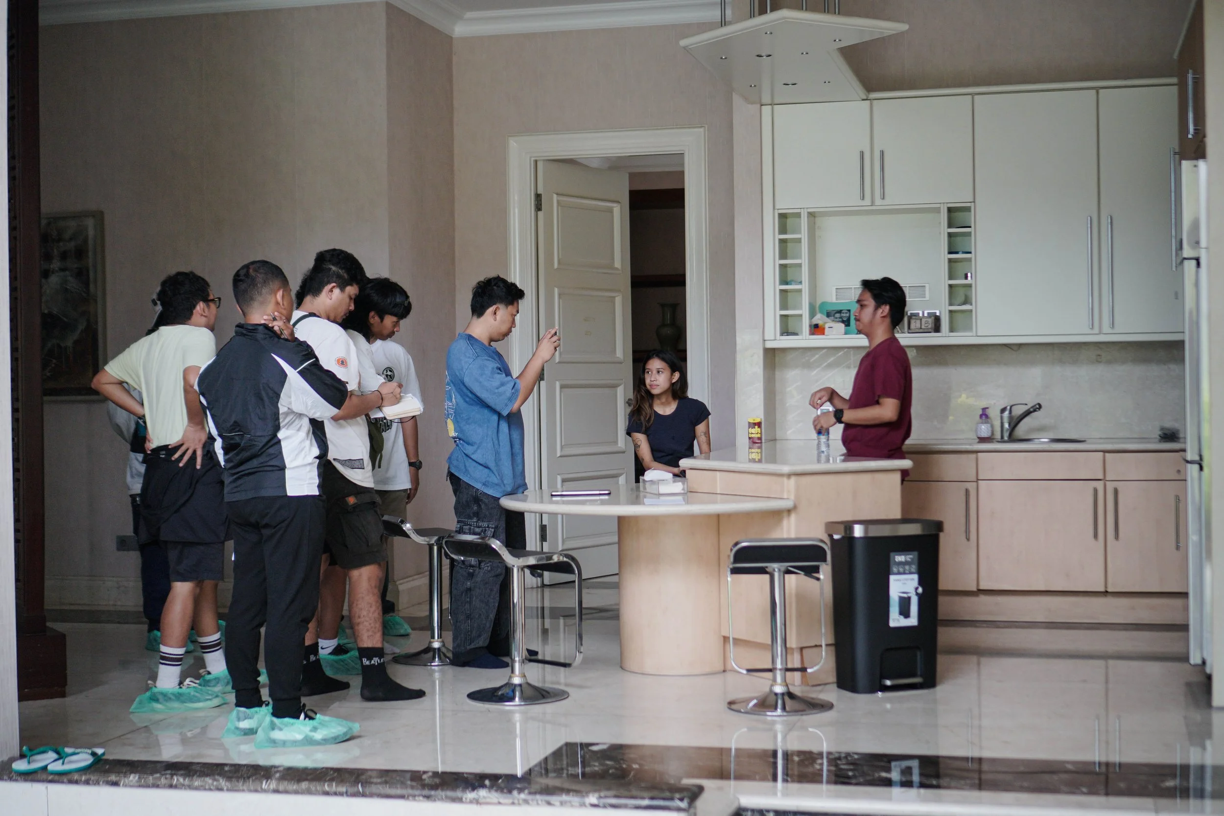 Group of young people standing in line inside a kitchen, with some wearing shoe covers, while two individuals are preparing food or drinks at the kitchen counter.