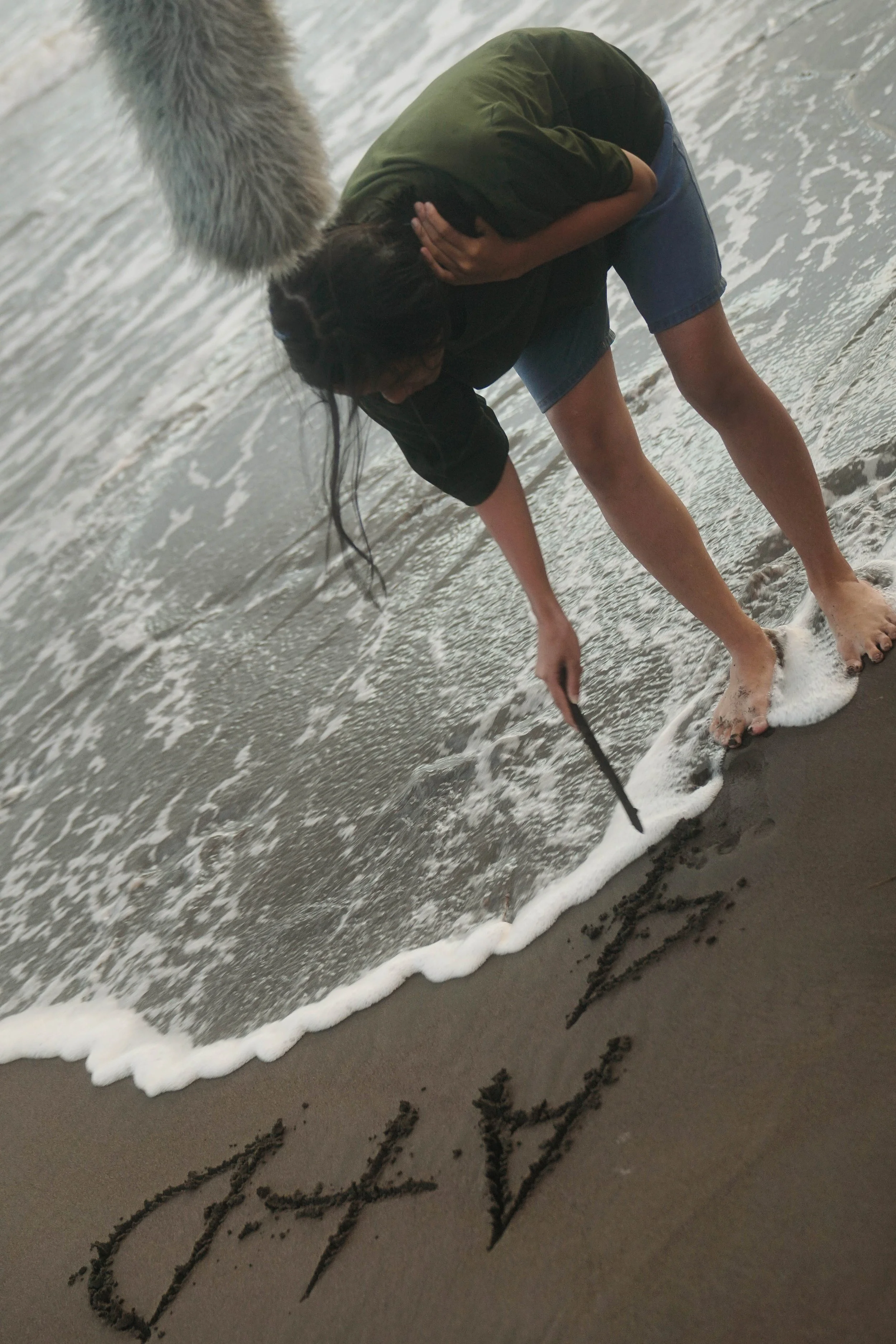 Person on ocean beach writing the word "LOVE" in the sand with a stick, while holding their head with one hand and wearing a green shirt and denim shorts.