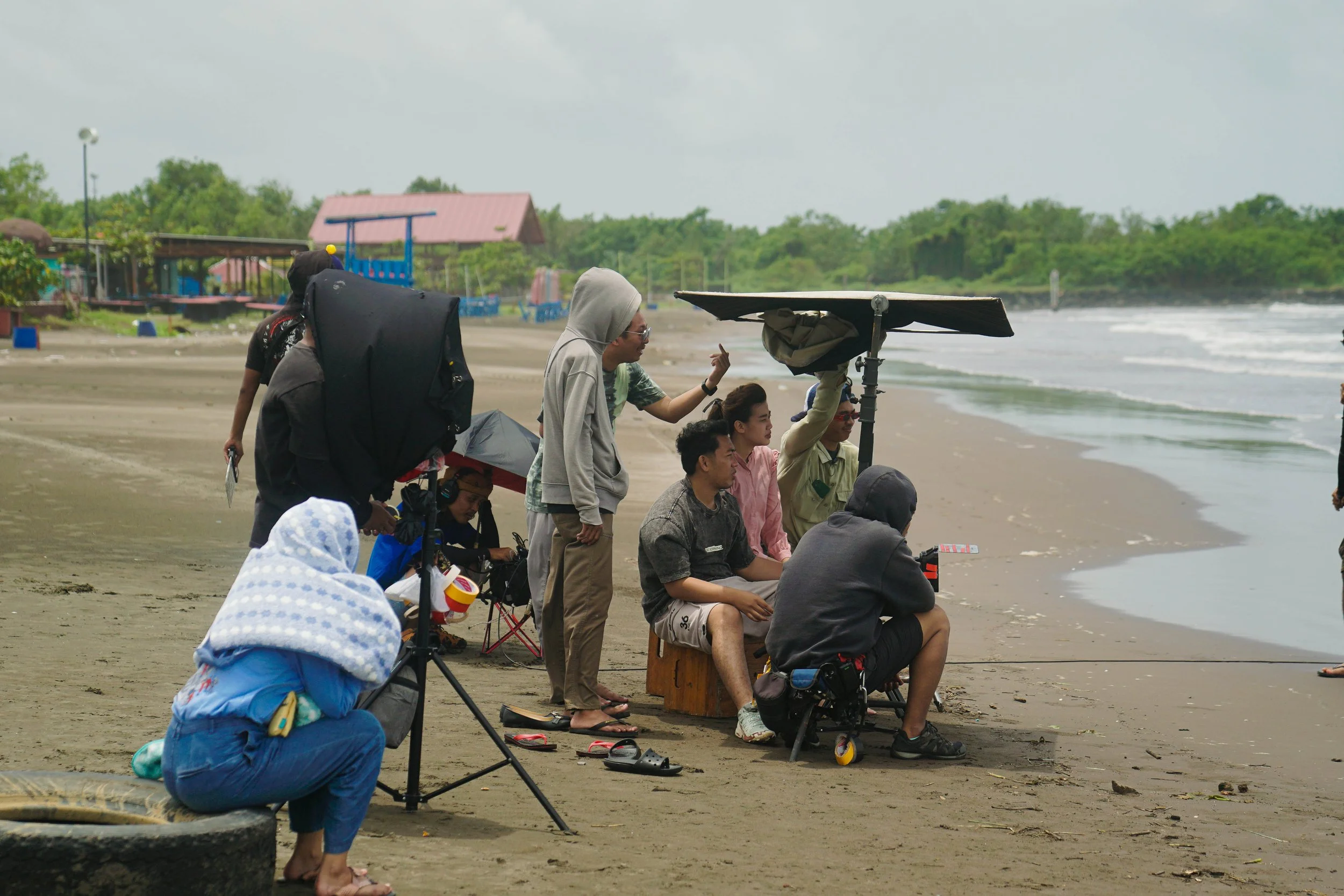 A group of people filming a scene on a beach with portable equipment, some sitting and others standing, with the ocean and a small building in the background.