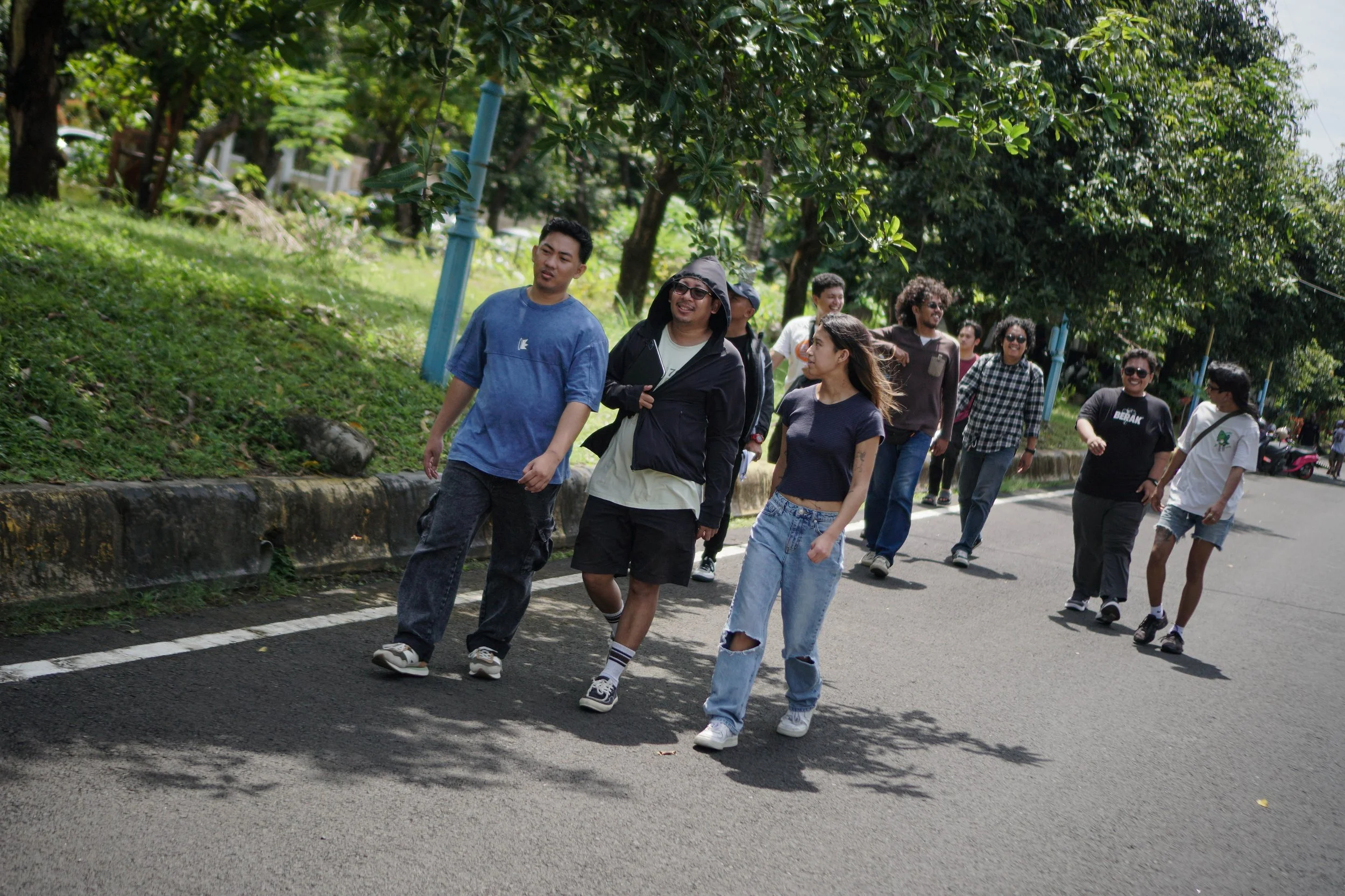 A group of young adults walking along a street with green trees and grass on the side, enjoying a sunny day.