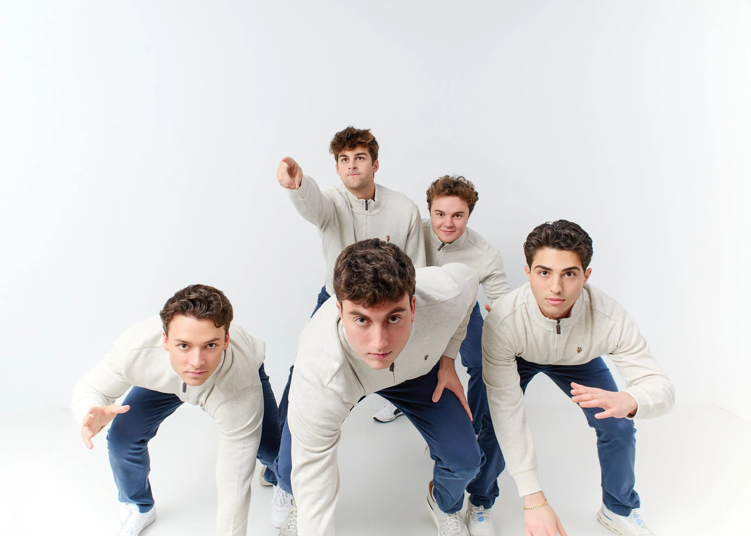 Five young men in matching beige zip-up sweatshirts and blue pants, posing in a race start position against a plain white background.