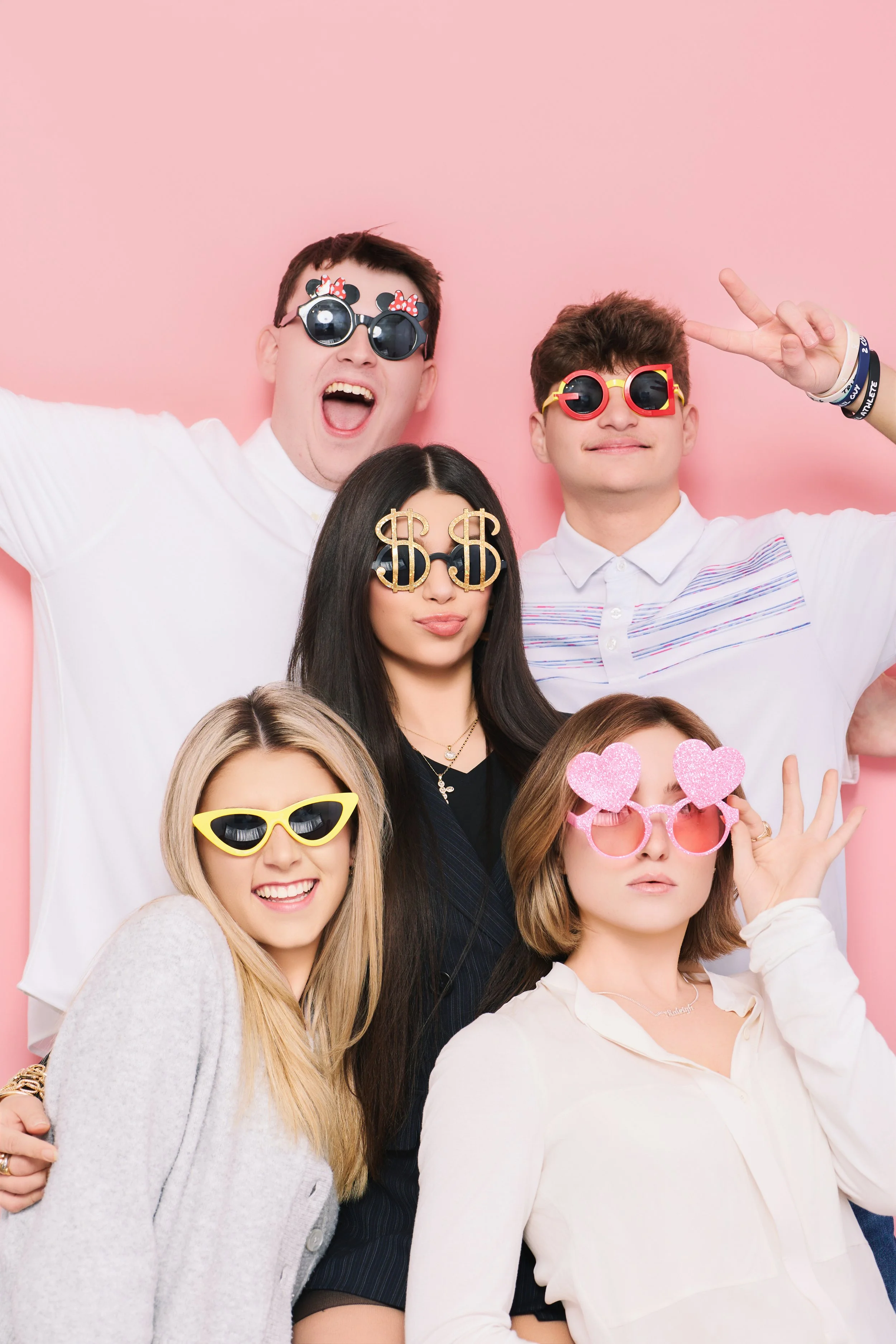 Group of five young adults wearing fun, colorful sunglasses against a pink background, posing for a photo.