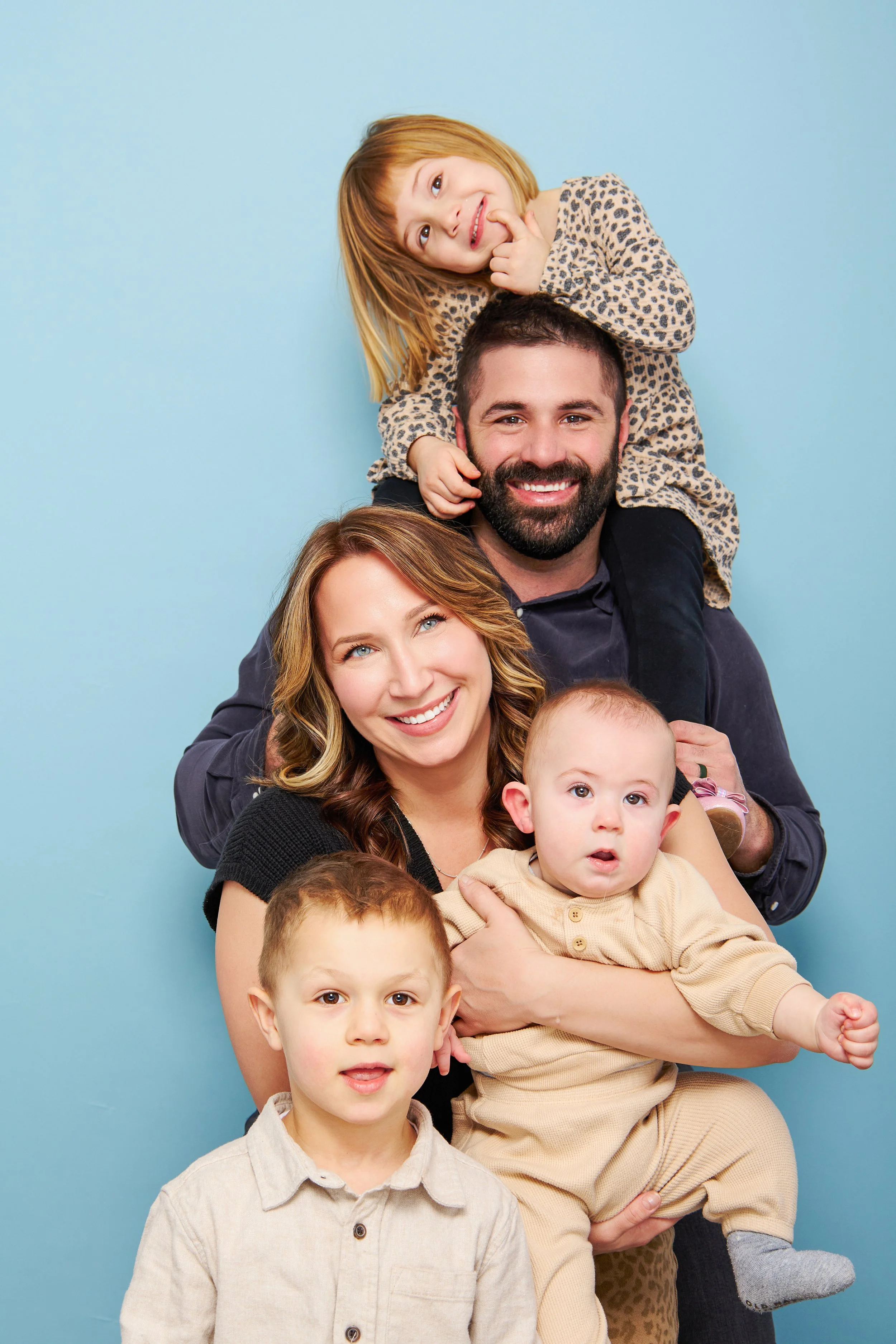 A happy family of five posing together against a light blue background. The father has dark hair and a beard, holding a young girl on his shoulders. The mother has wavy brown hair, holding a baby boy. A young boy with short light hair stands in front.