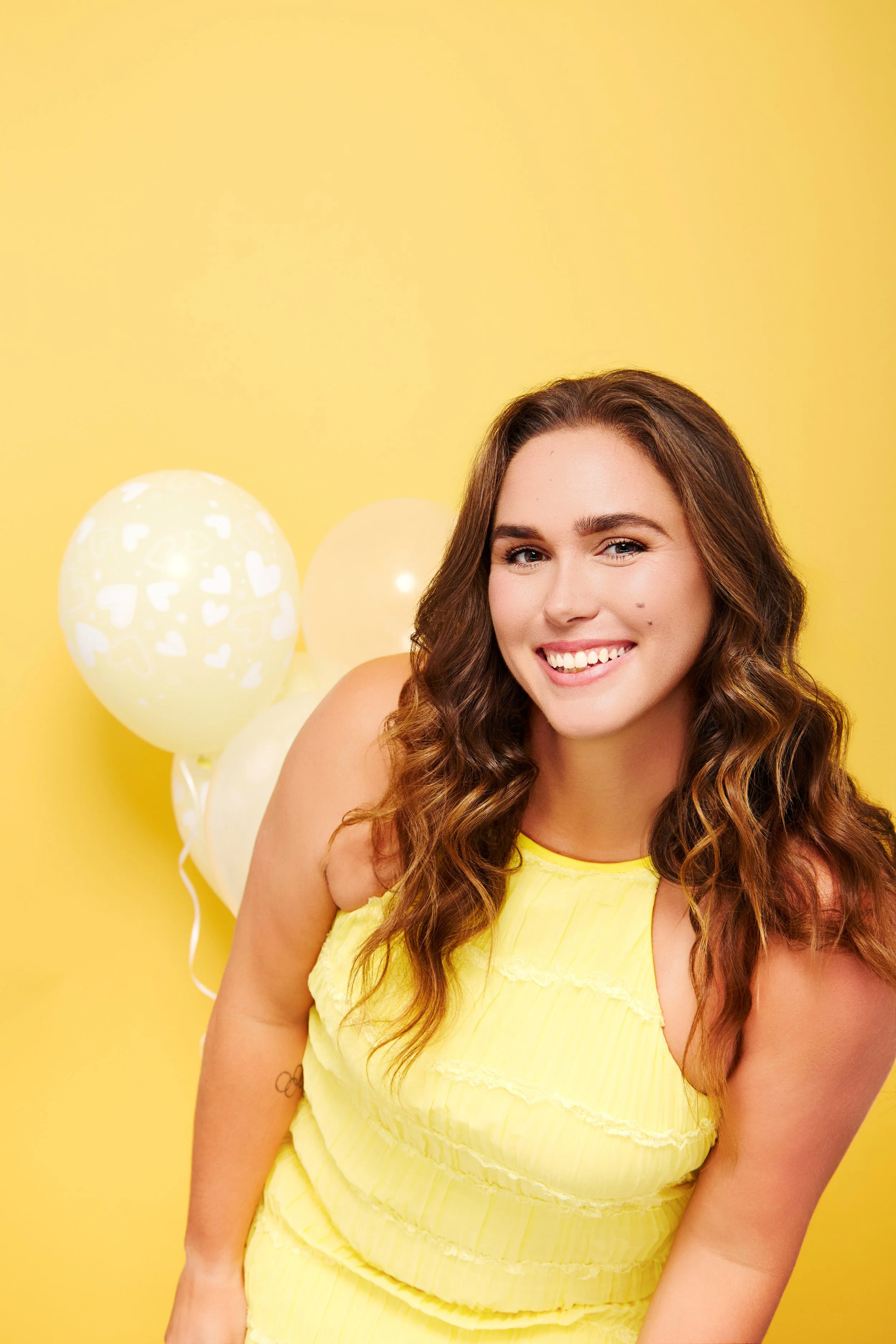 Young woman with wavy brown hair smiling in front of yellow background with white balloons.