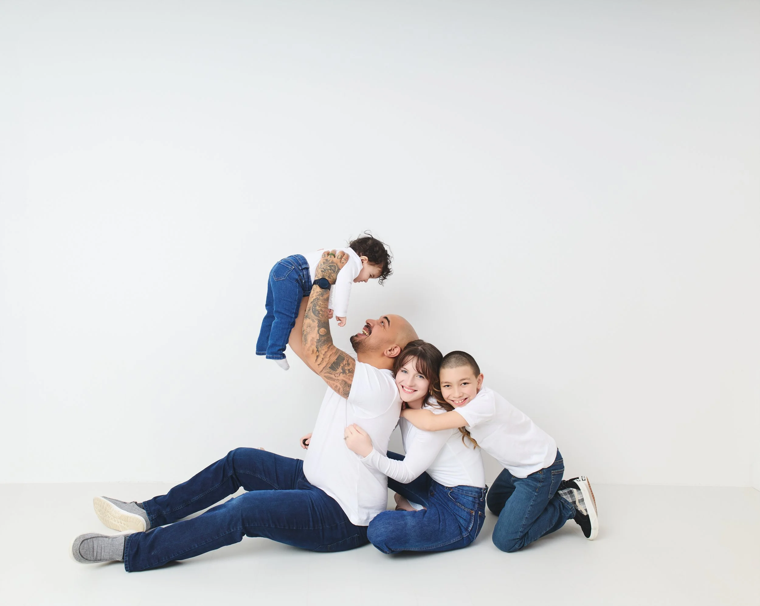 A family playing together against a plain white wall, with a father lifting a baby, and the mother and another child hugging and smiling on the floor.
