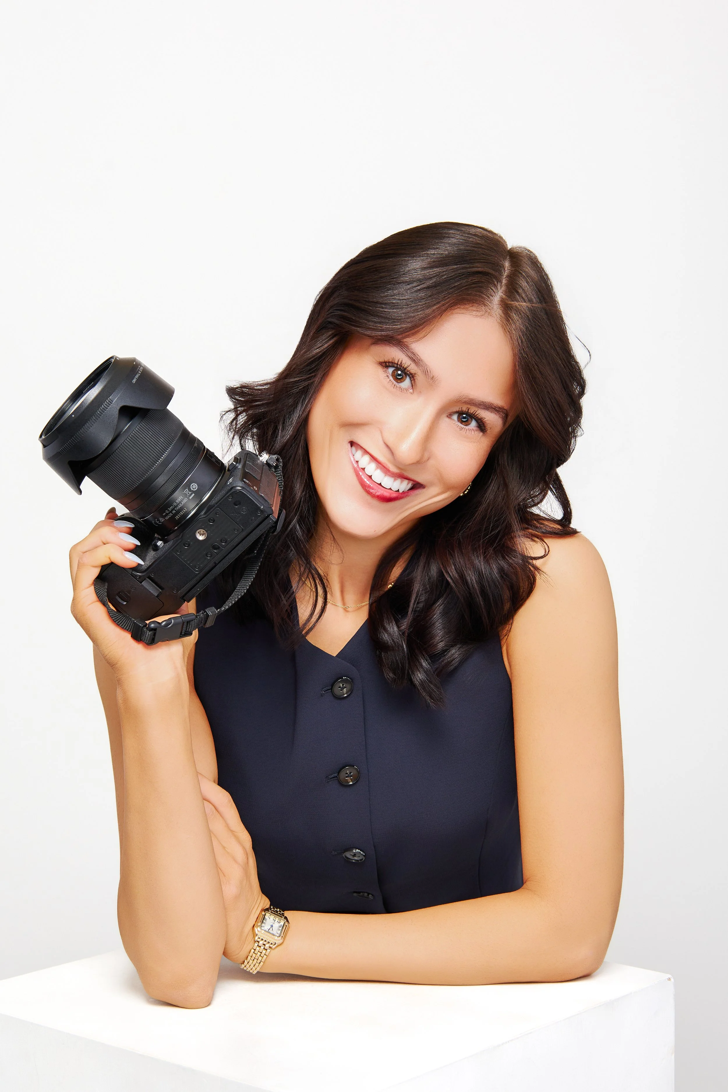 A woman with dark hair holding a camera, smiling, wearing a sleeveless navy blue top and a gold watch.