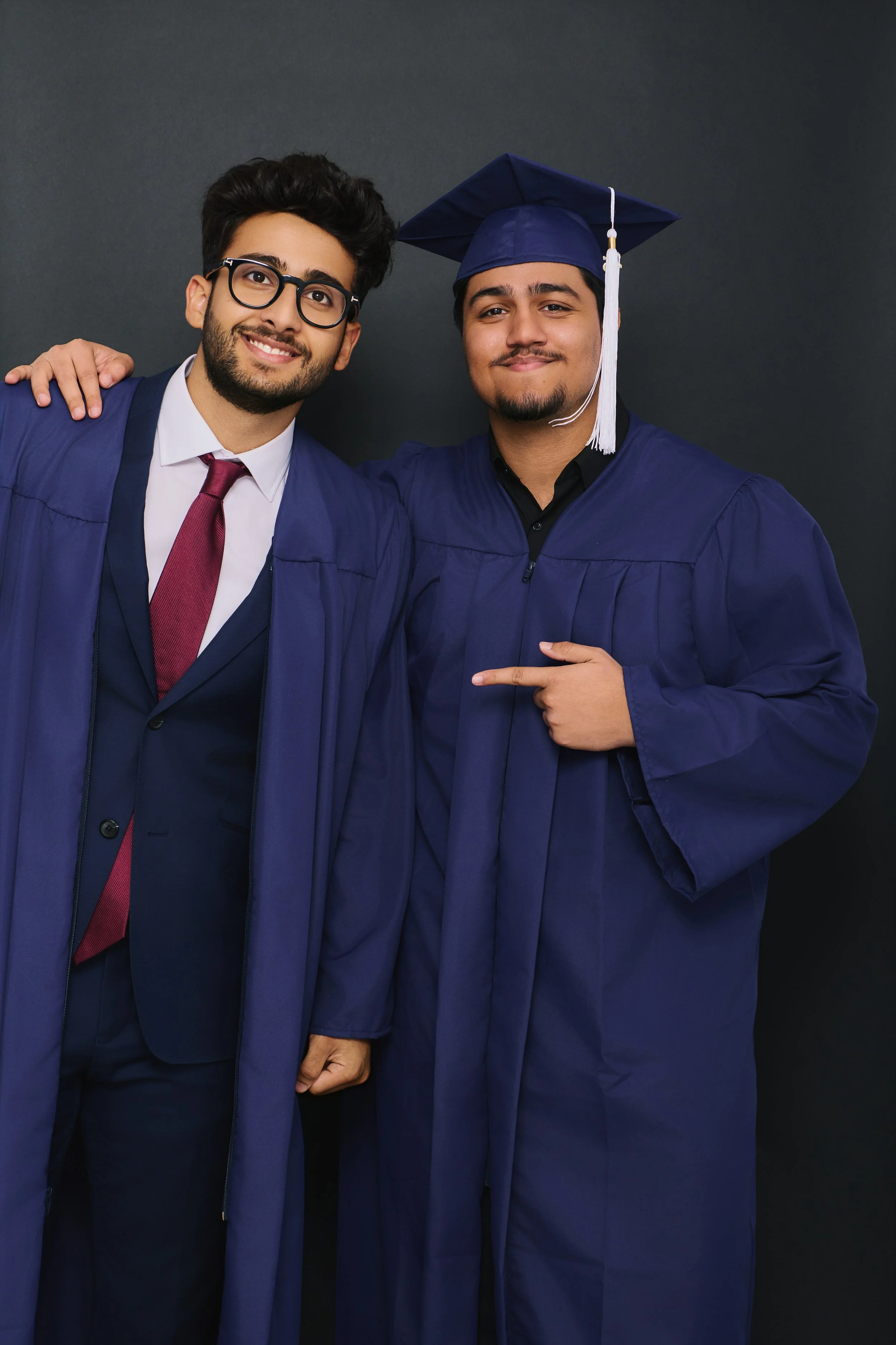 Two young men in graduation gowns and caps smiling, one with glasses and a tie, the other pointing at his cap, against a dark background.
