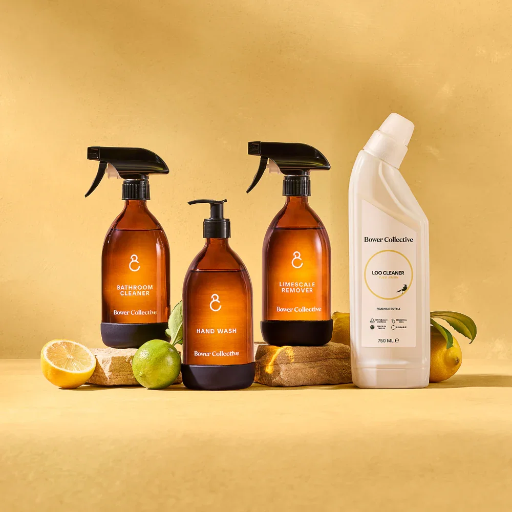 Bower Collective cleaning products including bathroom cleaner, hand wash, and limescale remover bottles on a yellow surface with lemons and limes around them.