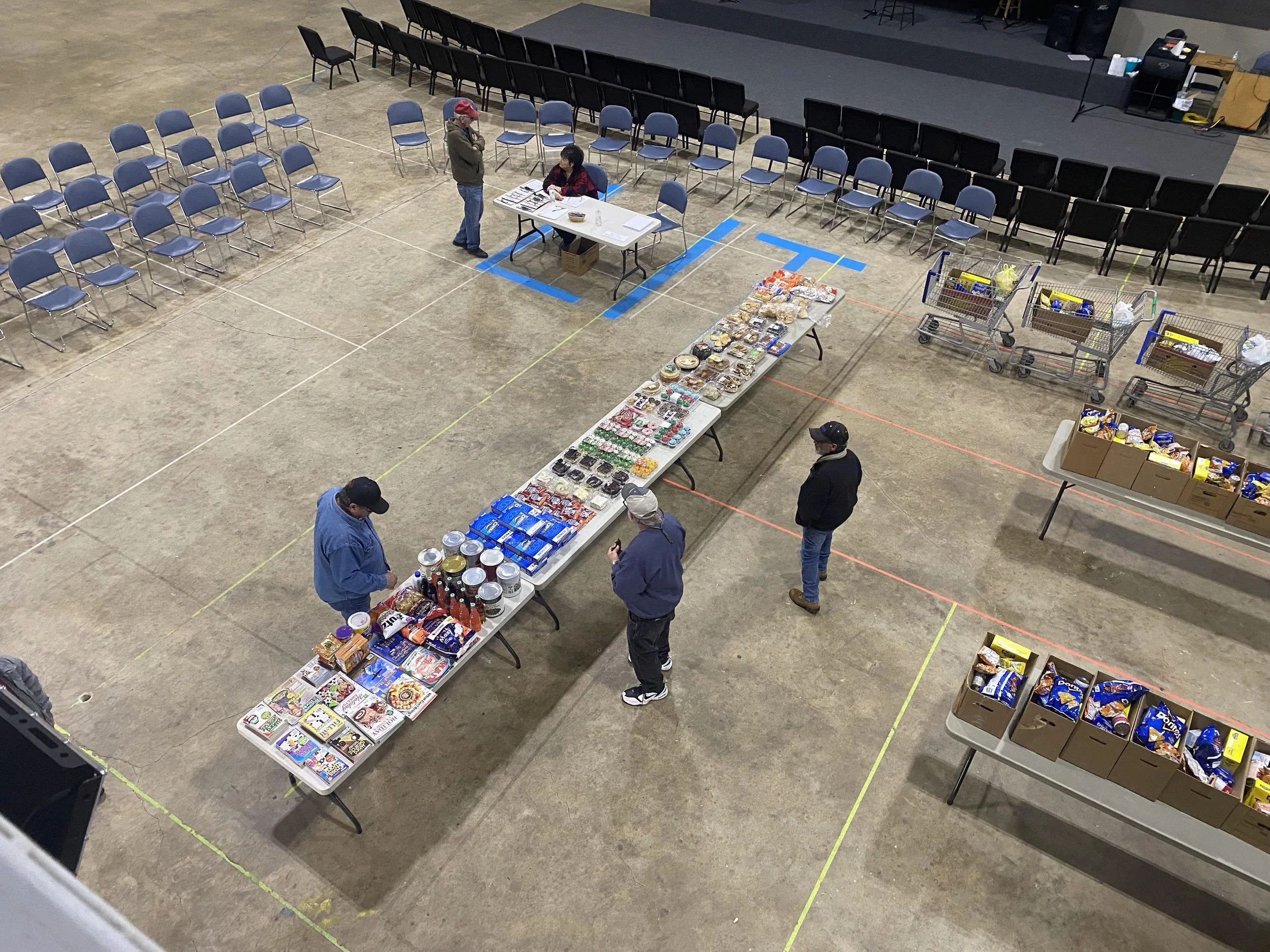 An indoor space set up for a food or goods distribution, with tables holding canned and packaged foods, shopping carts, and scattered chairs and boxes.