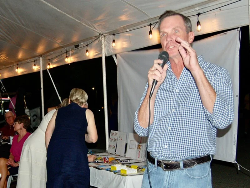 A man speaking into a microphone at an event under a tent, with several people seated around him and tables with displays.