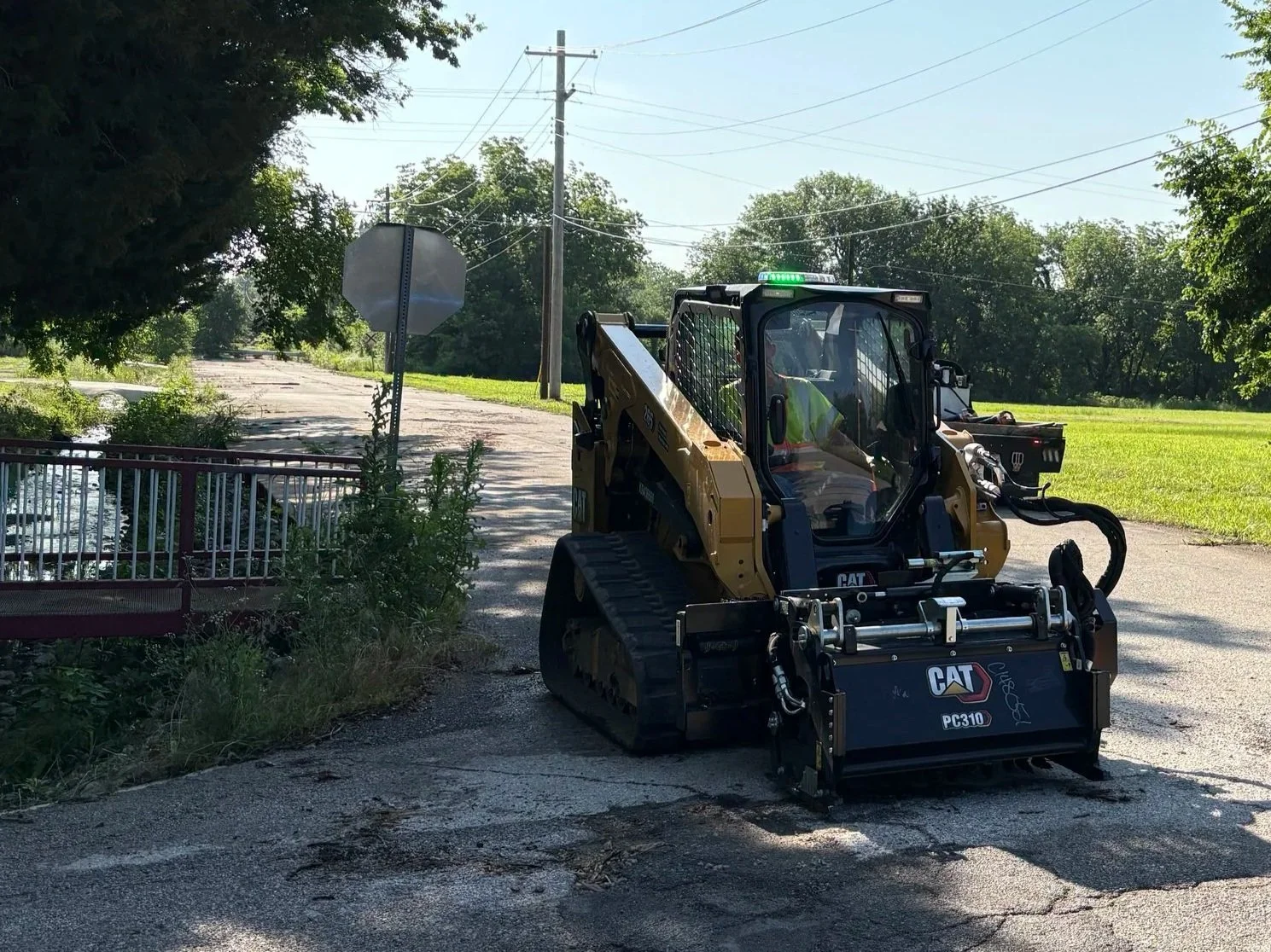 A small yellow Caterpillar skid steer loader working on a cracked asphalt surface near a grassy area, with a stop sign and utility poles in the background.
