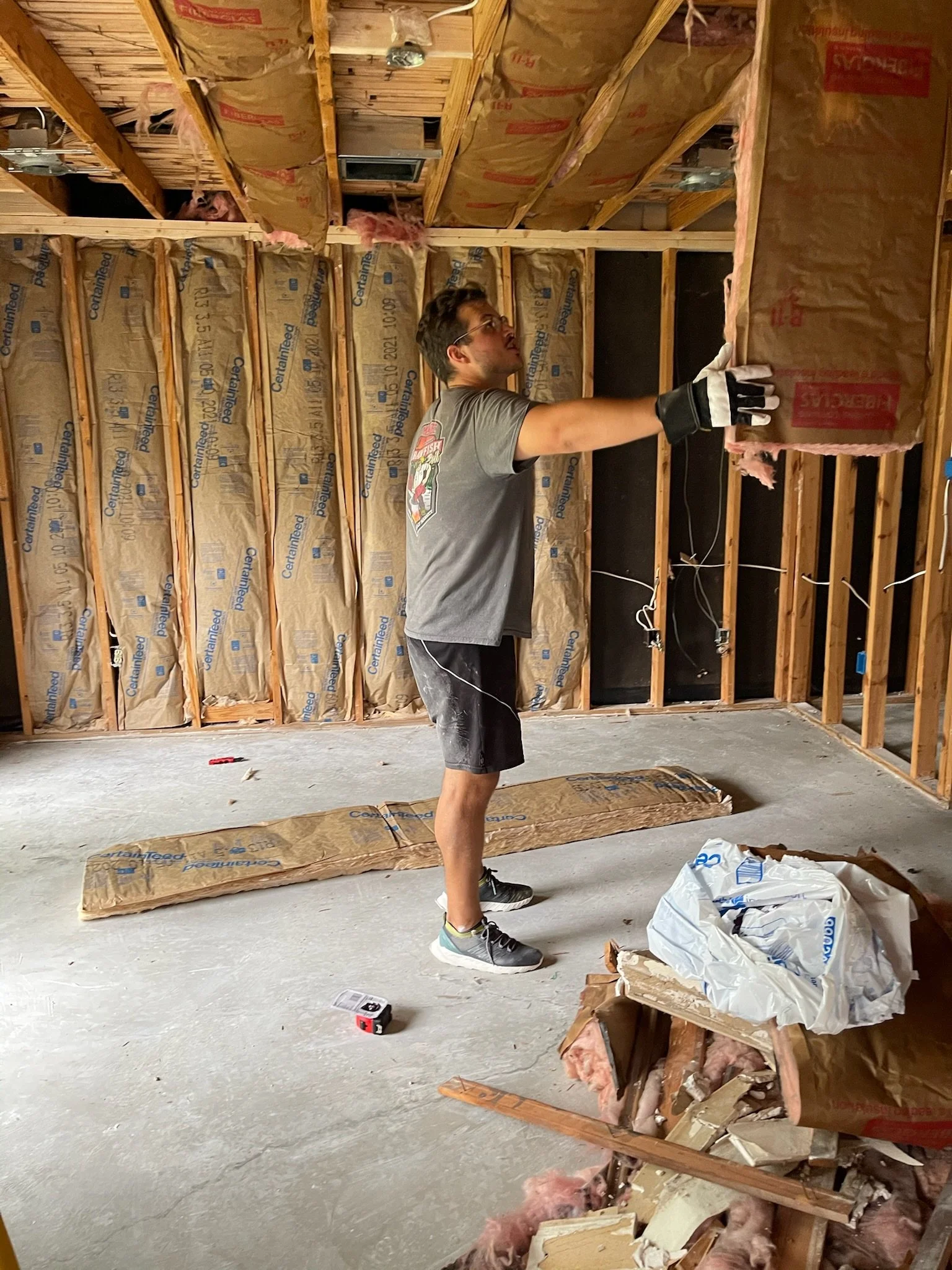 A man installing insulation in the walls of a house under construction. He is wearing gloves and casual clothes, standing on a concrete floor with unfinished wooden framing and insulation materials around him.