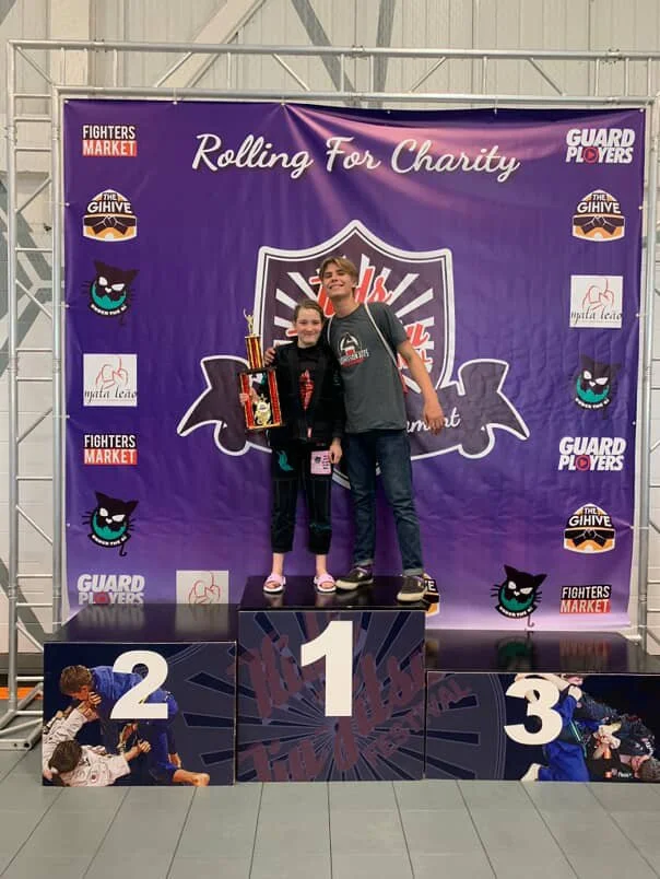 Two children standing on a winners' podium, the girl holding a trophy, at an awards event titled 'Rolling For Charity.' The boy is standing next to the girl, smiling, with a backdrop featuring logos and the phrase 'Rolling For Charity.' The podium levels are numbered 1, 2, and 3, with images of children in a martial arts setting on the sides of the lower levels.