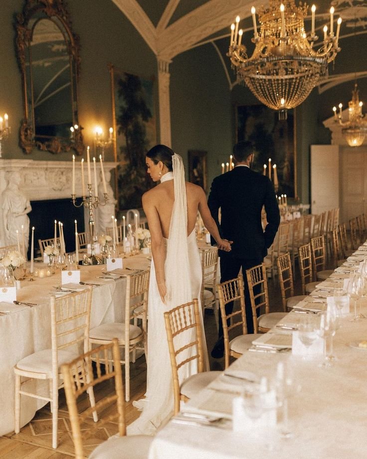 A bride in a white wedding gown and veil holding hands with a groom in a dark suit, walking through a decorated banquet hall with a long table, chandeliers, candelabras, and marble sculptures.