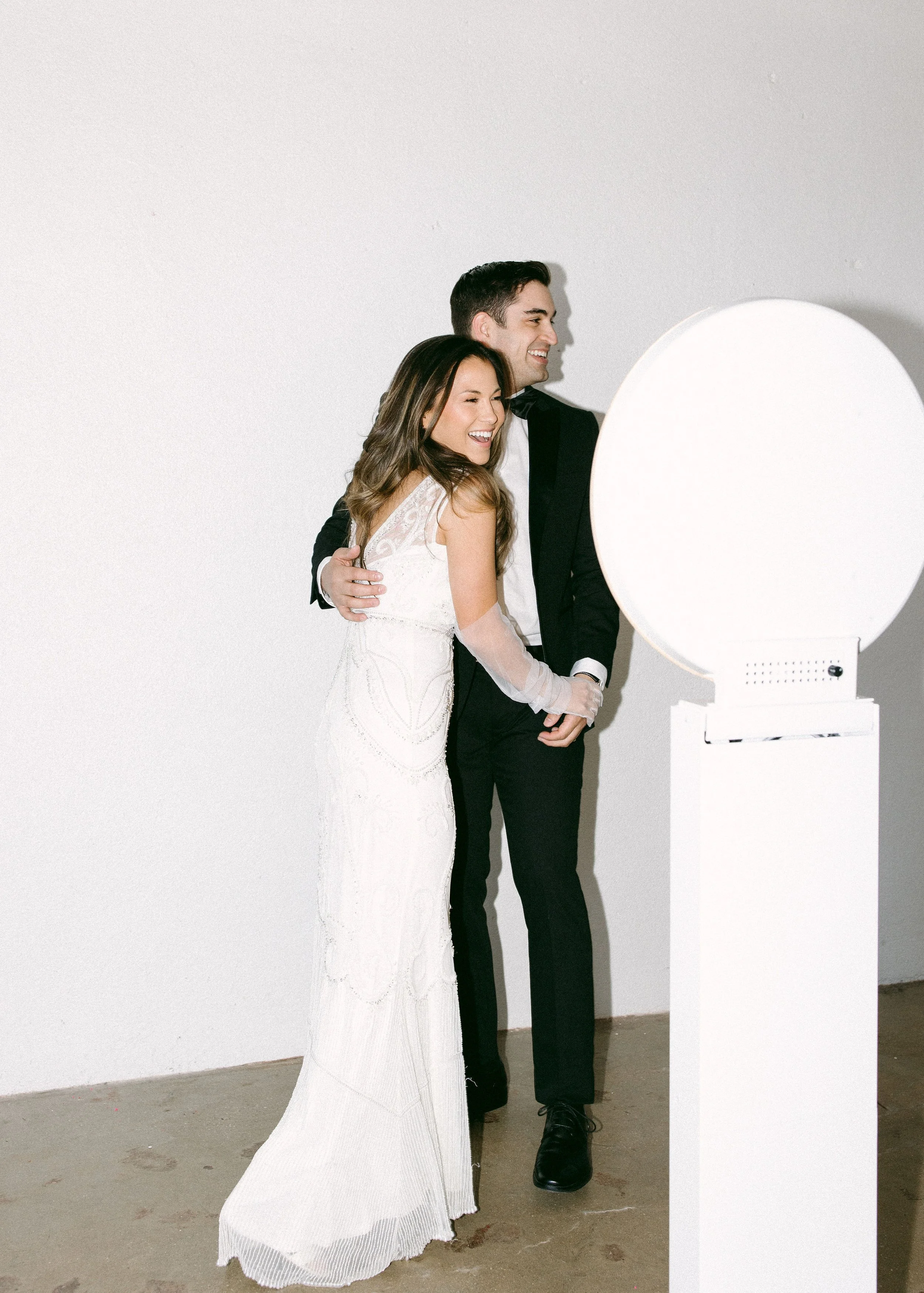 A smiling couple, dressed in formal wedding attire, shares a hug while looking at a large, illuminated mirror or photo stand in an art gallery or studio. The woman wears a white wedding dress, and the man wears a black tuxedo.
