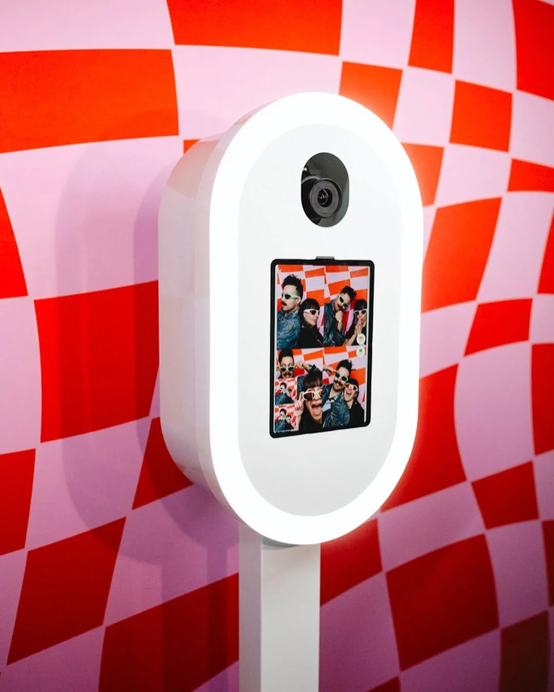 Photo of a photo booth with a camera at the top, a screen displaying a group of four people taking selfies, and a colorful red and pink checkered background.