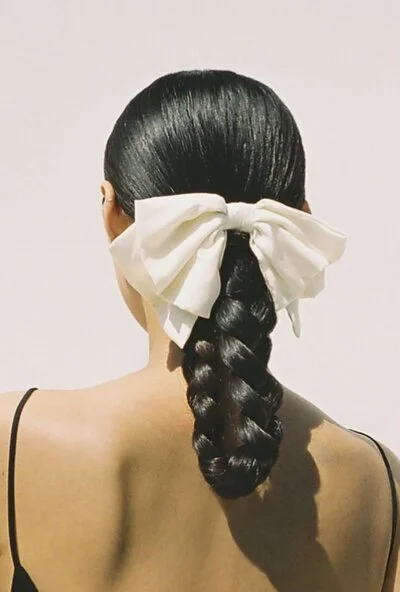 Back view of a woman with long black hair styled in a braid secured with a large white bow.
