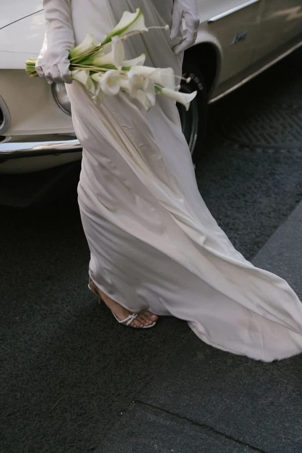 A person in a white dress holding a bouquet of white calla lilies, standing near a classic beige or tan car on a street.