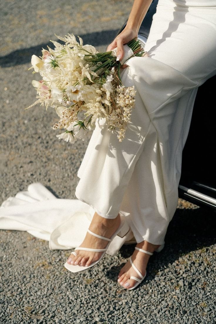 Close-up of a bride wearing white heels and holding a bouquet of white and light pink flowers, with a white wedding dress on a gravel surface.