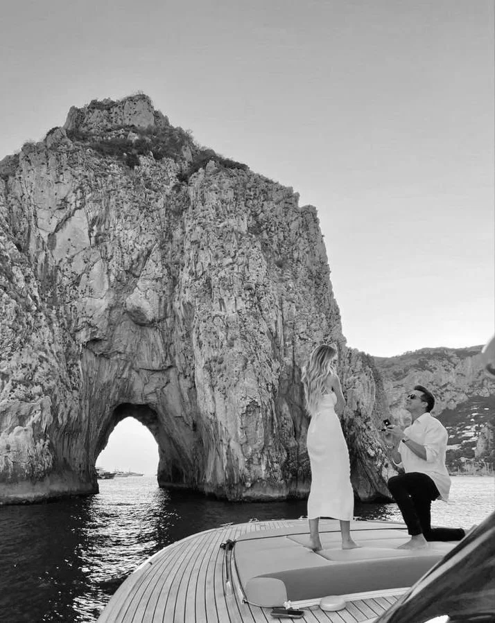 A man proposing marriage to a woman on a boat near a large rocky arch formation in the water.