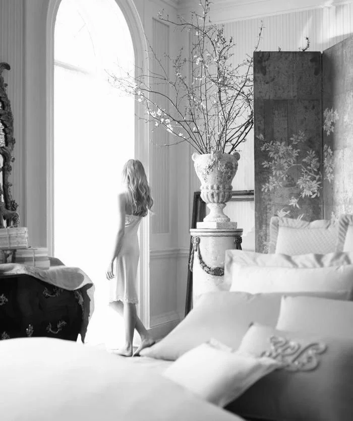 A woman with long hair standing by a window with sheer curtains, looking outside in a decorated bedroom with a large vase holding branches, an ornate dresser, and plush bedding.