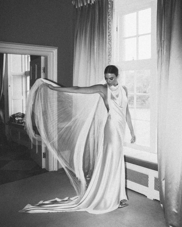A woman in a long, elegant white dress standing indoors next to a large window, holding a flowing part of her dress with one arm extended.