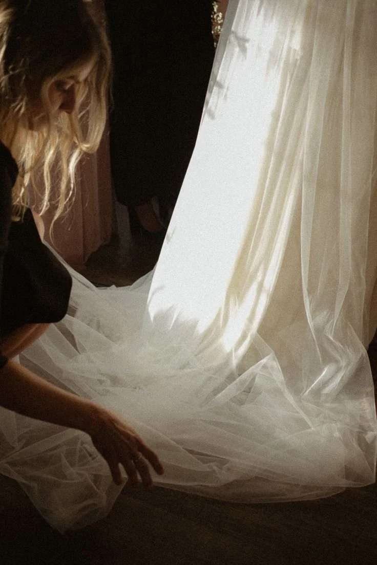 A woman with curly blonde hair is arranging a long, white, sheer fabric curtain near a window with bright sunlight shining through.