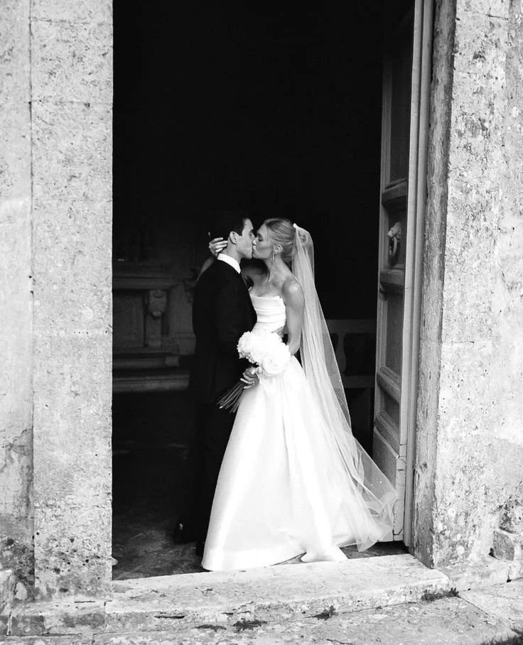A black-and-white photo of a bride and groom kissing in a doorway, with the bride holding a bouquet of flowers.